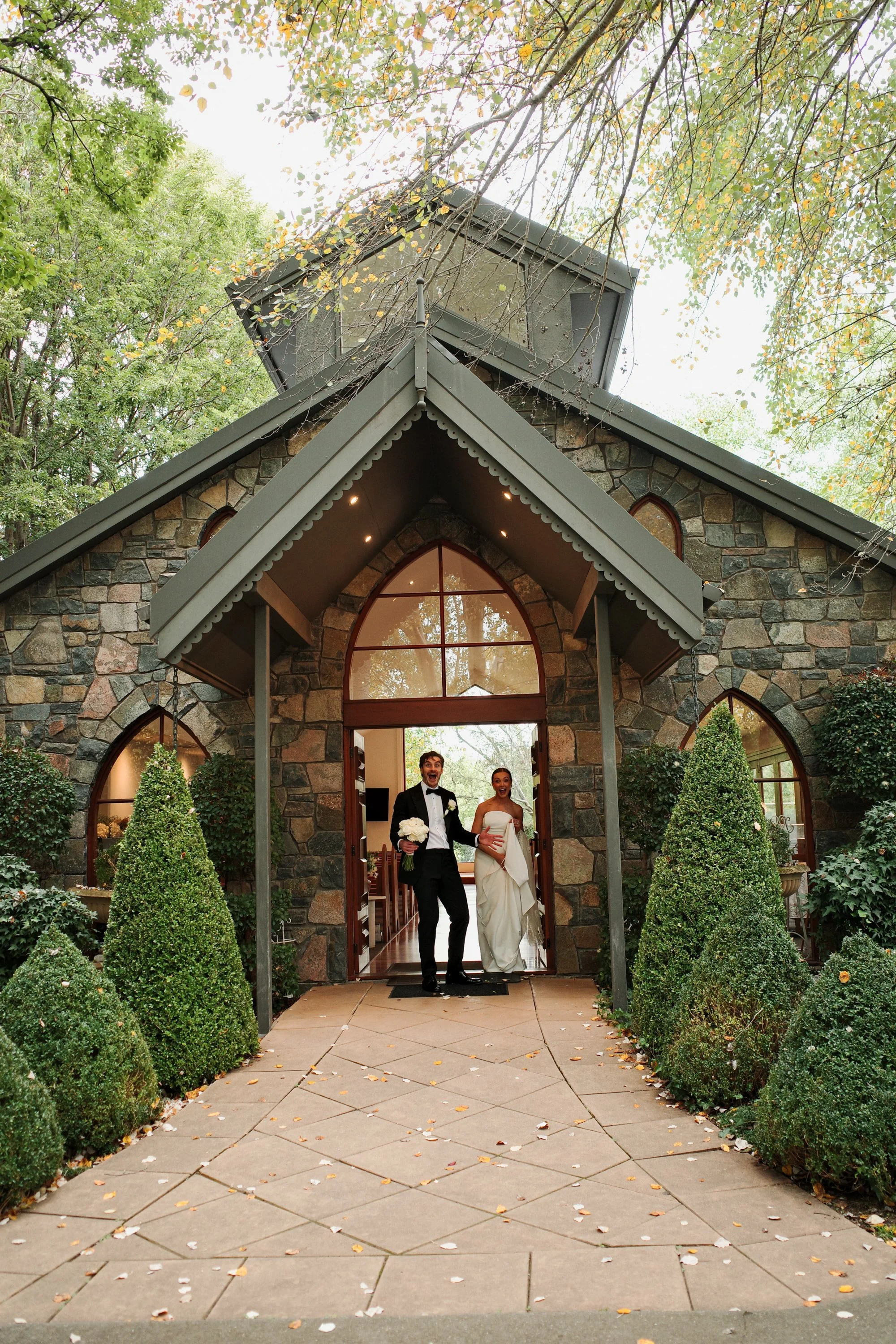 A newly married couple exiting a stone church on a cloudy day, surrounded by bushes and trees with some leaves on the ground, indicating fall. The groom is in a tuxedo holding a bouquet, and the bride is in a wedding dress with a veil, both smiling a
