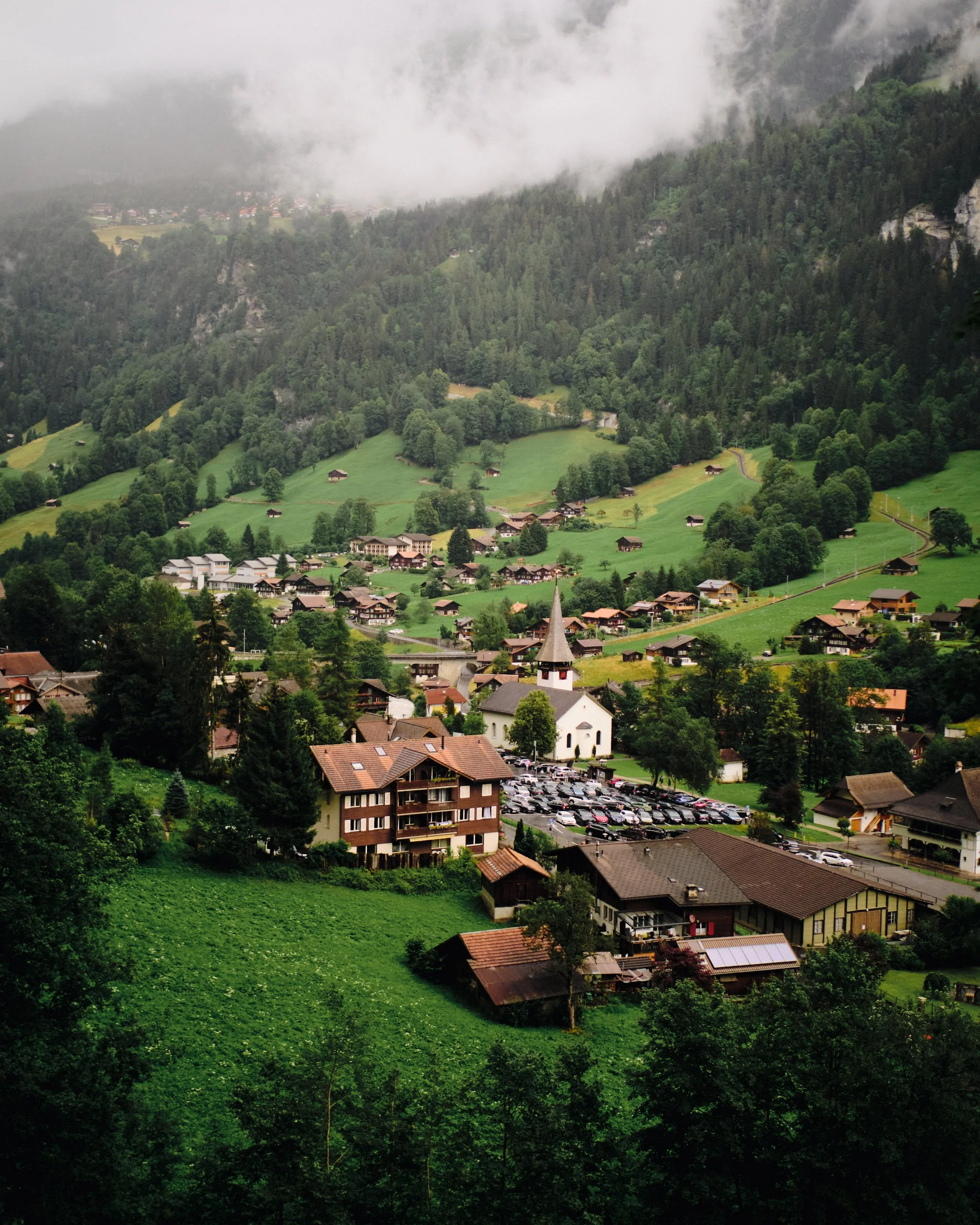 A lush green valley with a small village, scattered houses, and a church with a pointed steeple, surrounded by dense forested hills and mountains partially covered in low-hanging clouds.