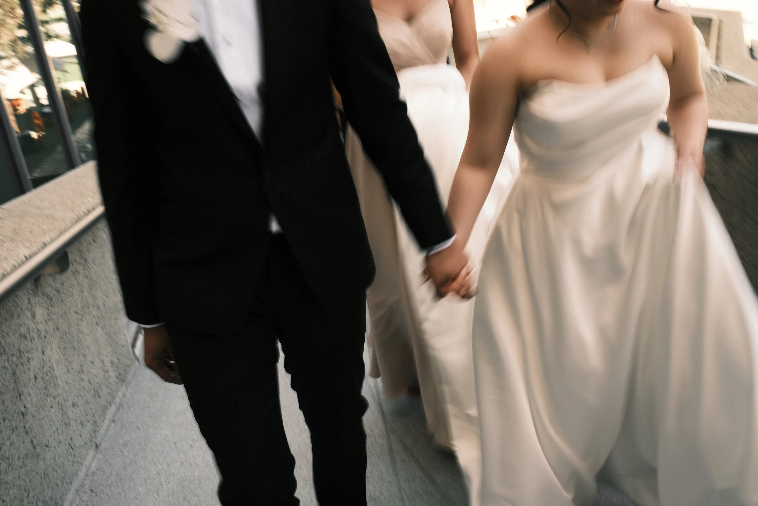 Close-up of a bride and groom holding hands, with the bride wearing a strapless white wedding gown and the groom in a black suit, walking together.