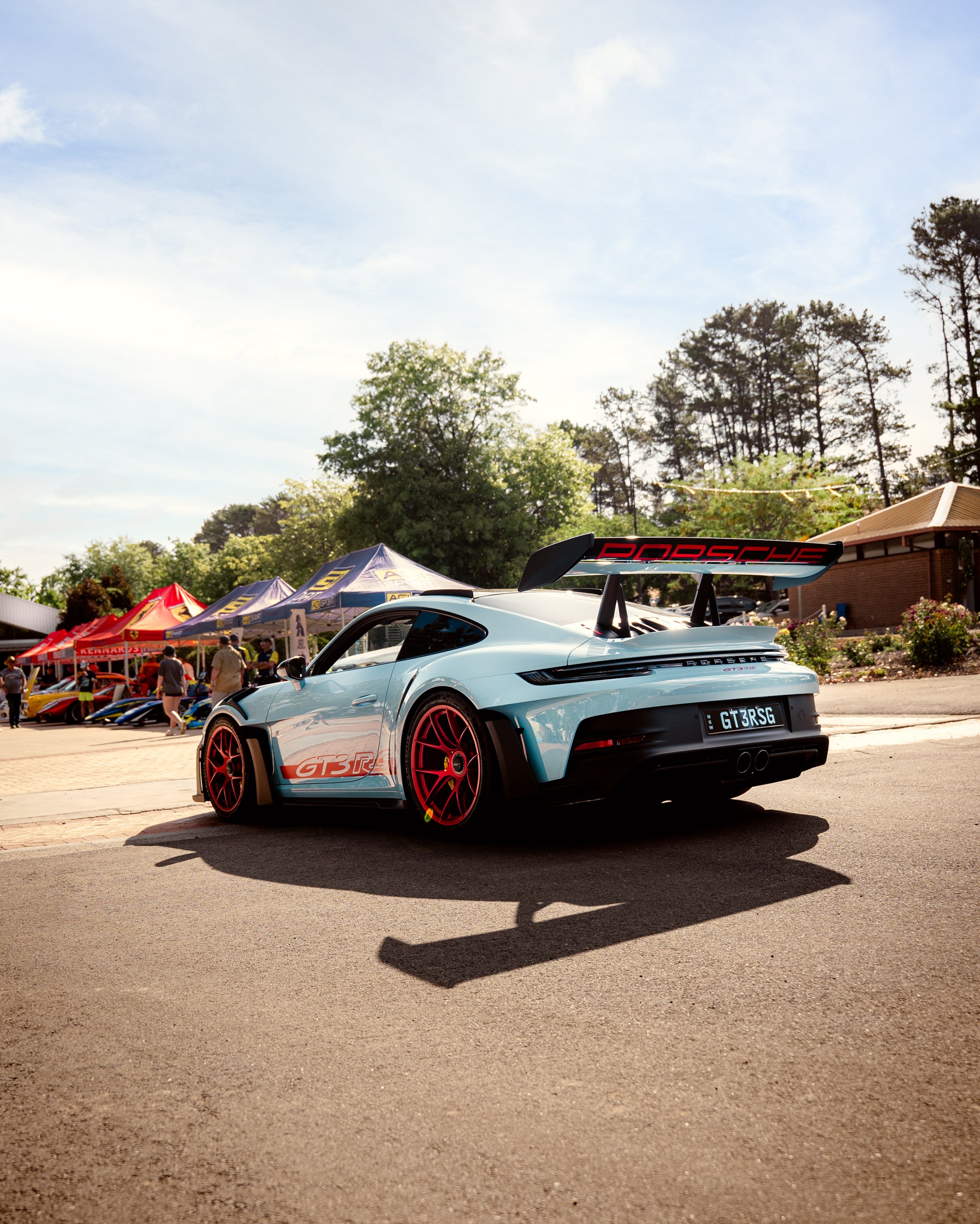 A white Porsche sports car with red accents and a large rear spoiler, parked at an outdoor gathering with tents and trees in the background.