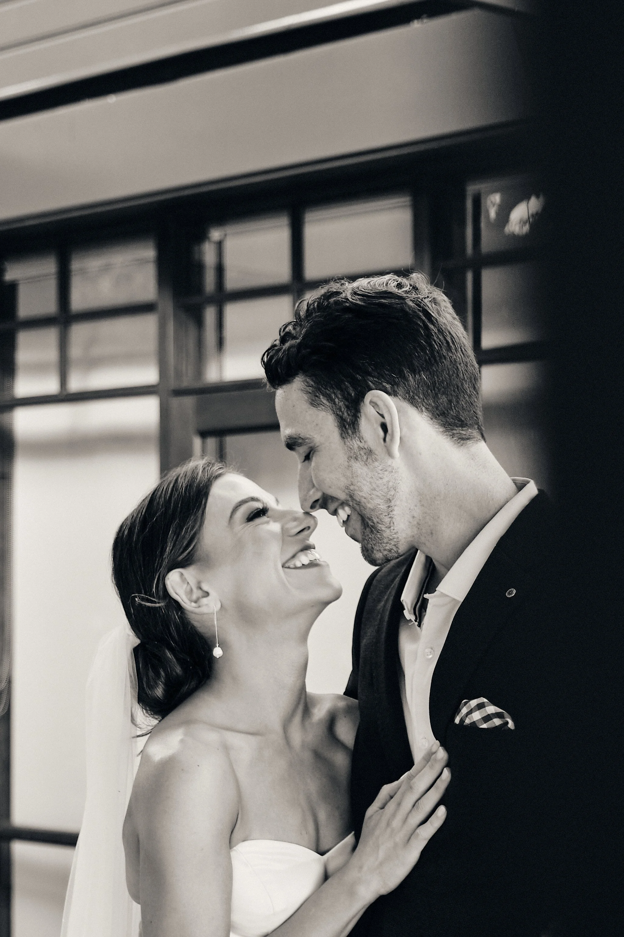 A bride and groom sharing a joyful moment at their wedding, with the bride smiling and the groom leaning in close, indoors with a background featuring a large window.