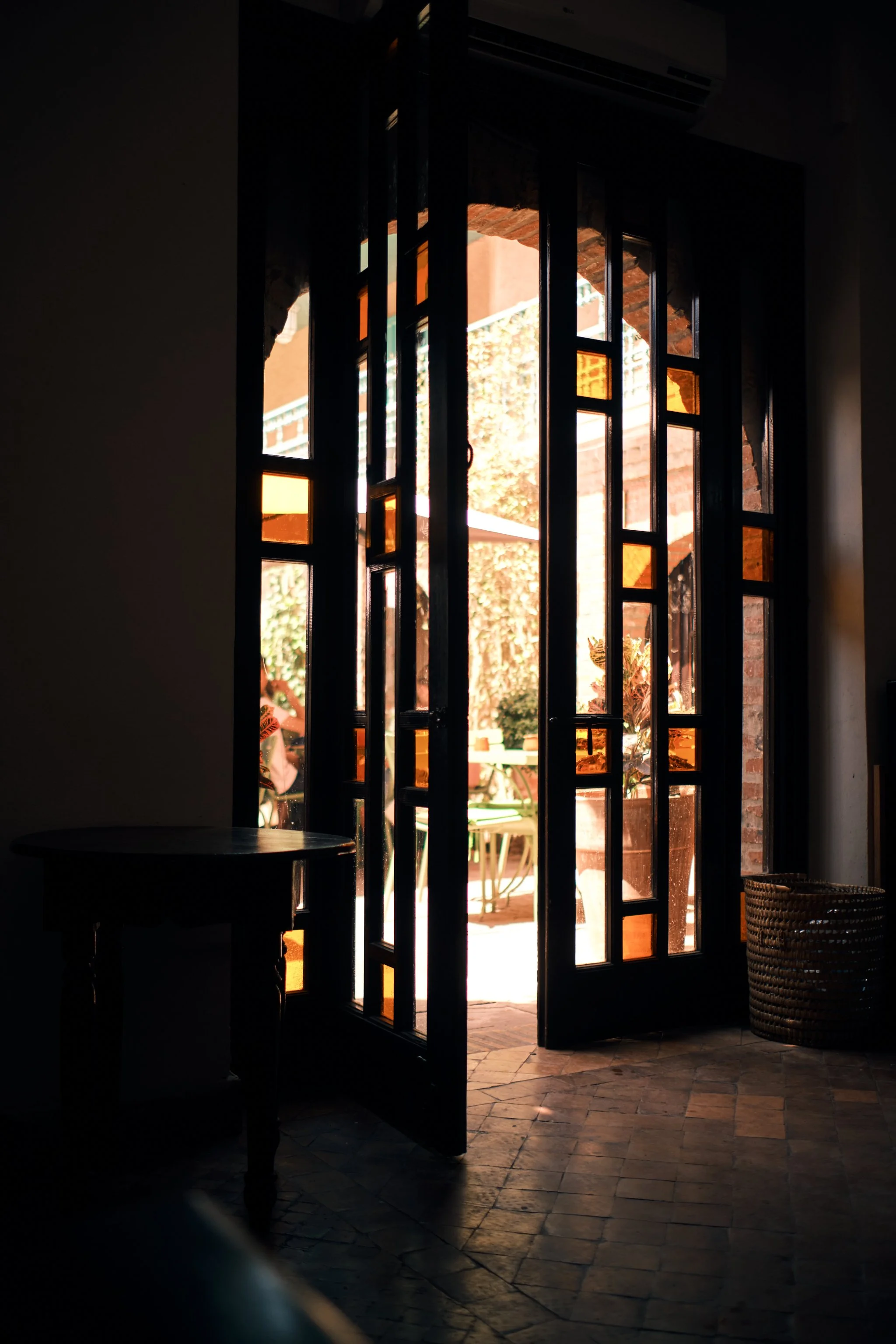Open glass door leading to a sunny outdoor patio with potted plants and outdoor furniture. Indoor space is dimly lit with a small table and a basket visible inside.