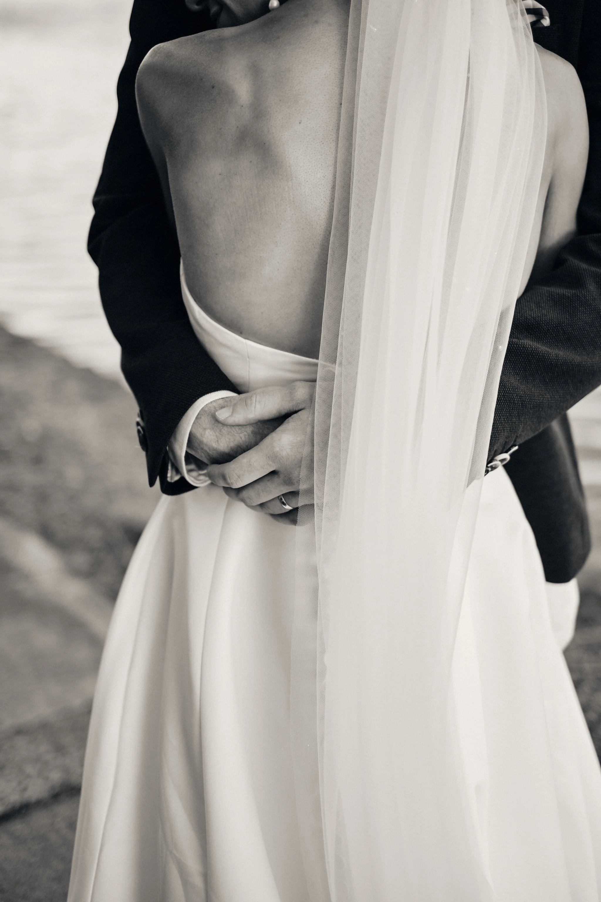 Close-up of a bride in a white wedding dress and veil, with a man in a dark suit standing behind her with his hands on her waist.