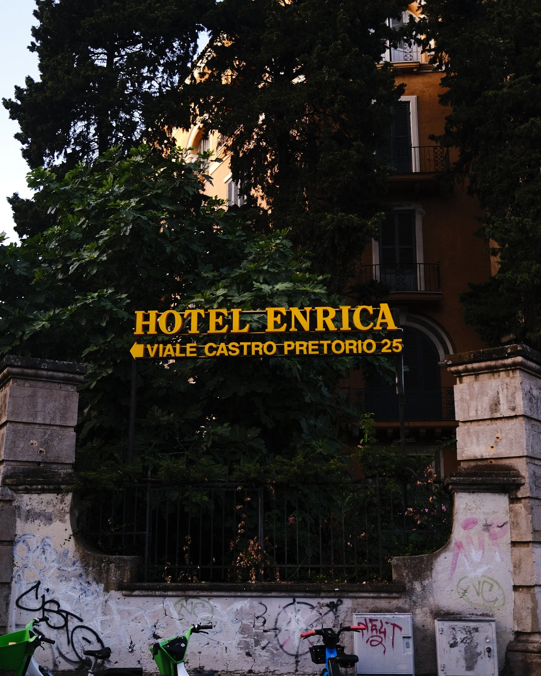 A street scene with a building partly hidden behind trees and a wall with graffiti. There are bicycles parked at the bottom of the wall and a sign indicating directions to Hotel Enrica and Viale Castro Pretorio 25.