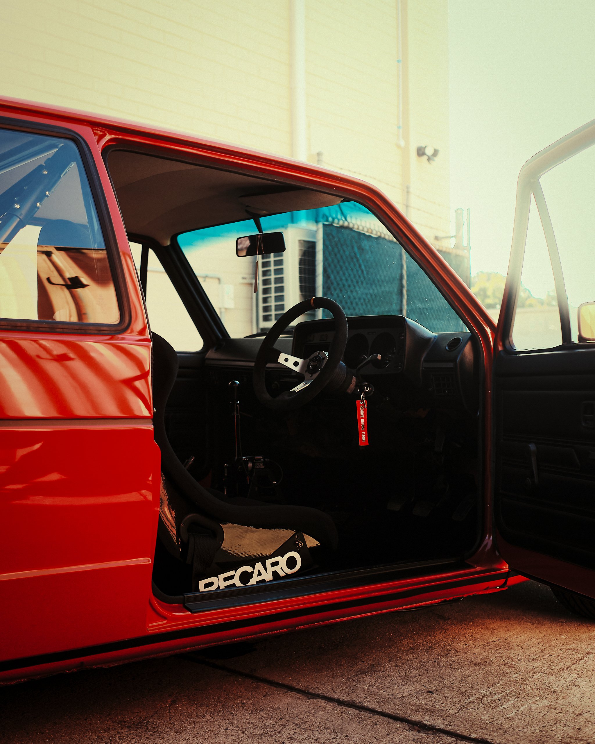 Red vehicle with an open door revealing the interior, including a steering wheel, dashboard, and a black seat, parked on a paved surface with a beige building and an air conditioning unit in the background.
