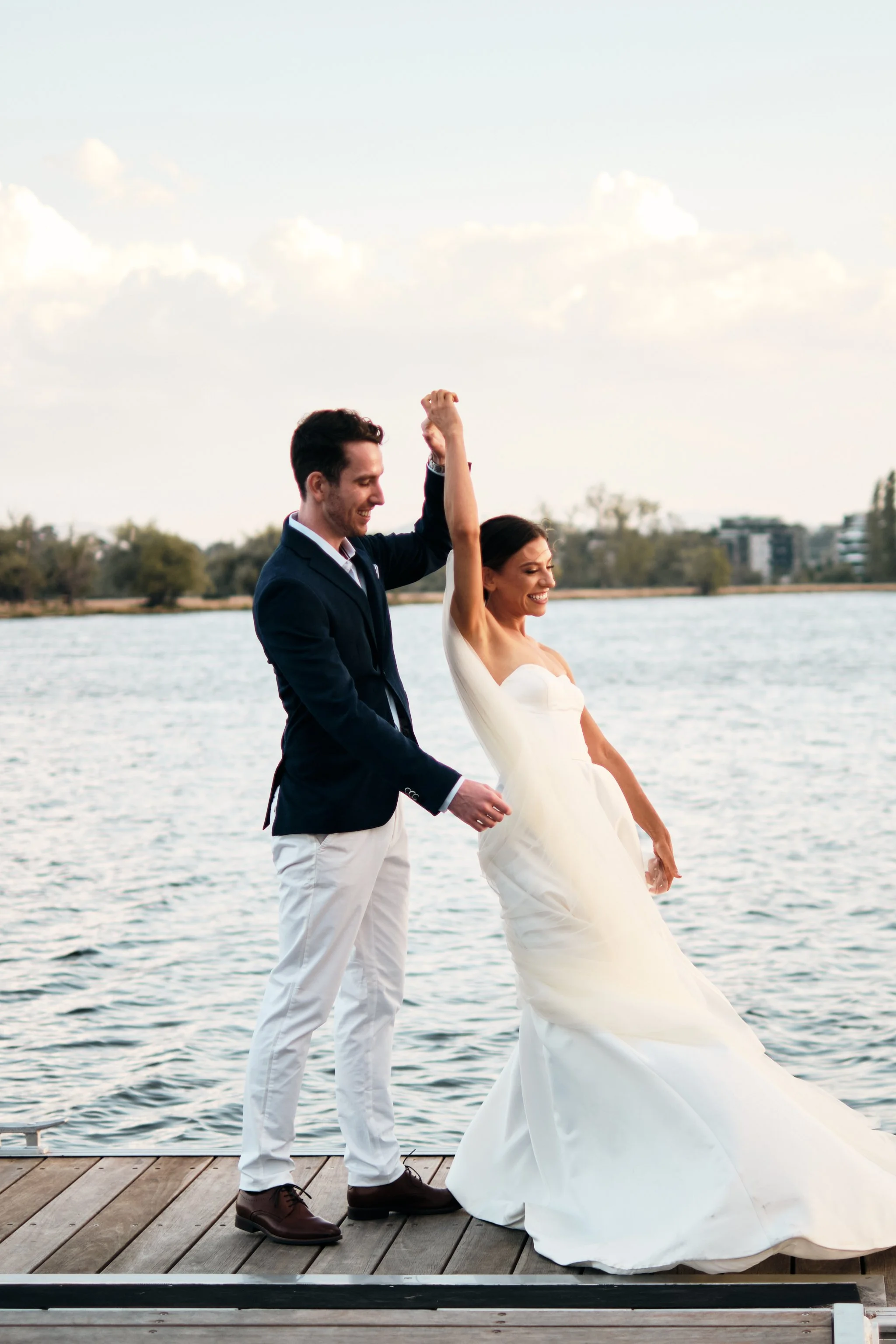 A man and woman celebrating on a wooden dock by a body of water. The man is dressed in a navy blazer and white pants, raising his arm in celebration. The woman is in a white wedding dress, smiling with her eyes closed as they share a joyful moment.