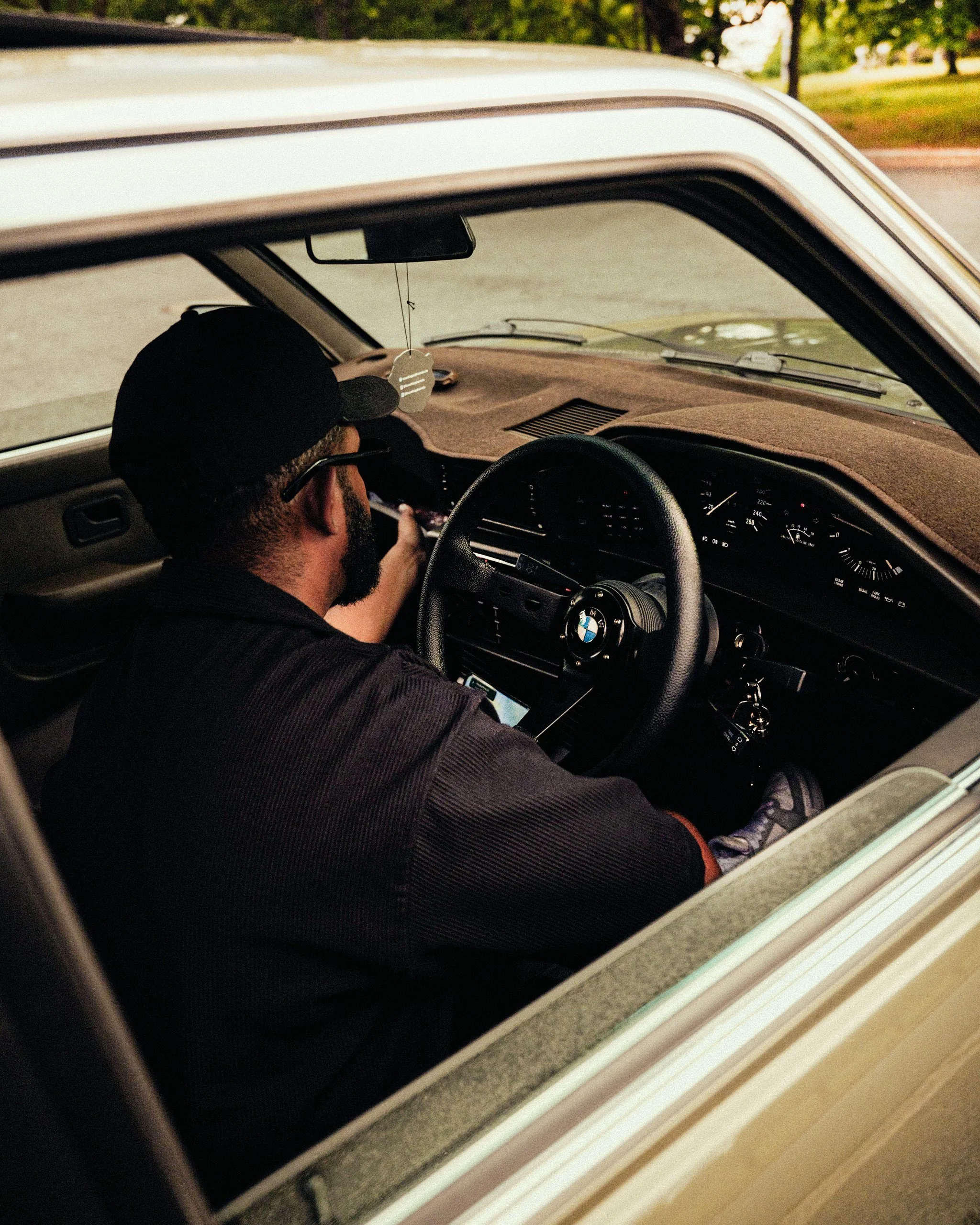 A man with a beard and wearing sunglasses and a black cap sitting in the driver's seat of a car, looking at a smartphone. The photo is taken from outside the car through the open window.
