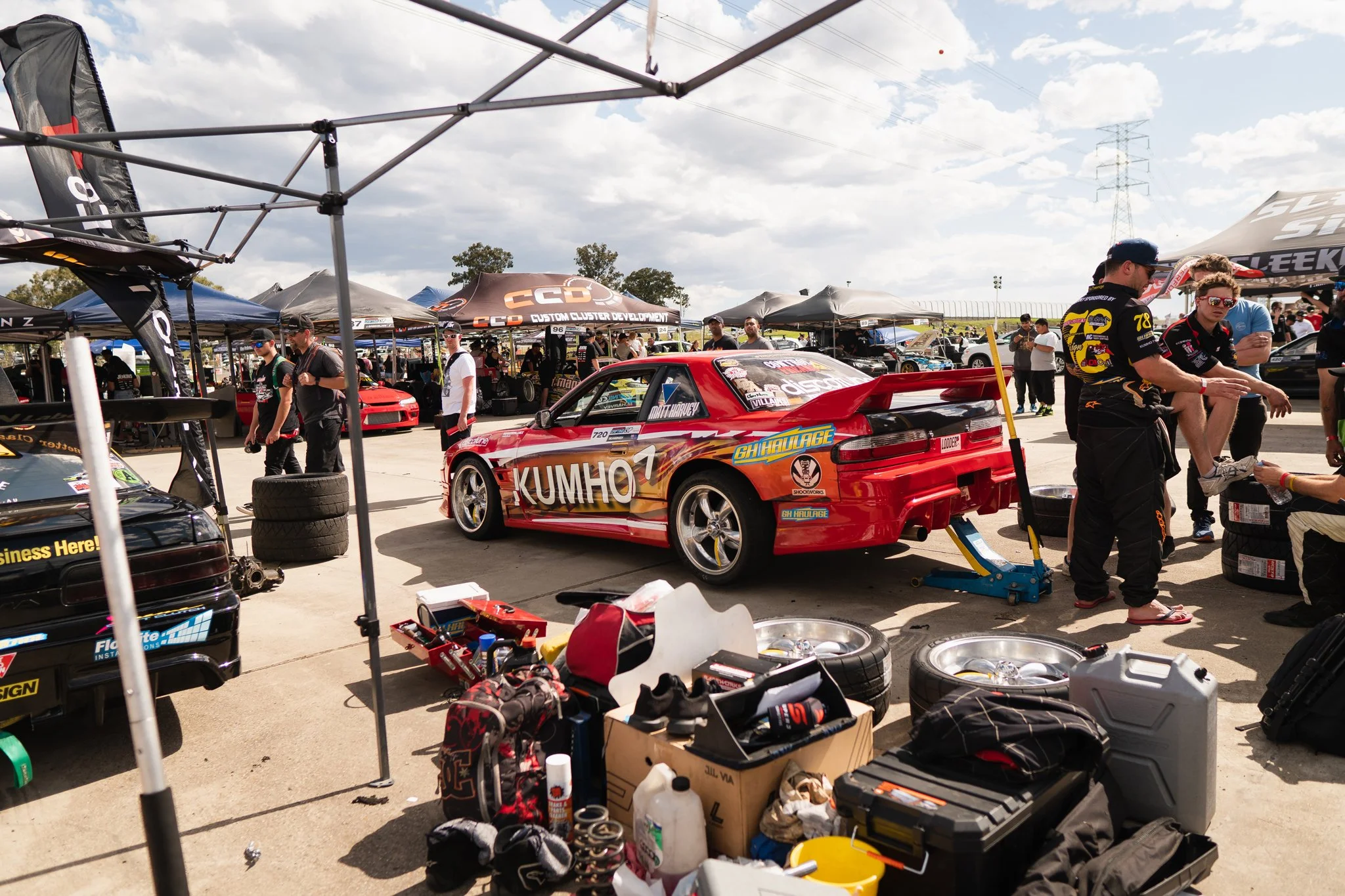 A busy motorsport paddock area with a red race car on a lift, surrounded by mechanics, team members, and equipment, with various tents and more race cars in the background on a partly cloudy day.