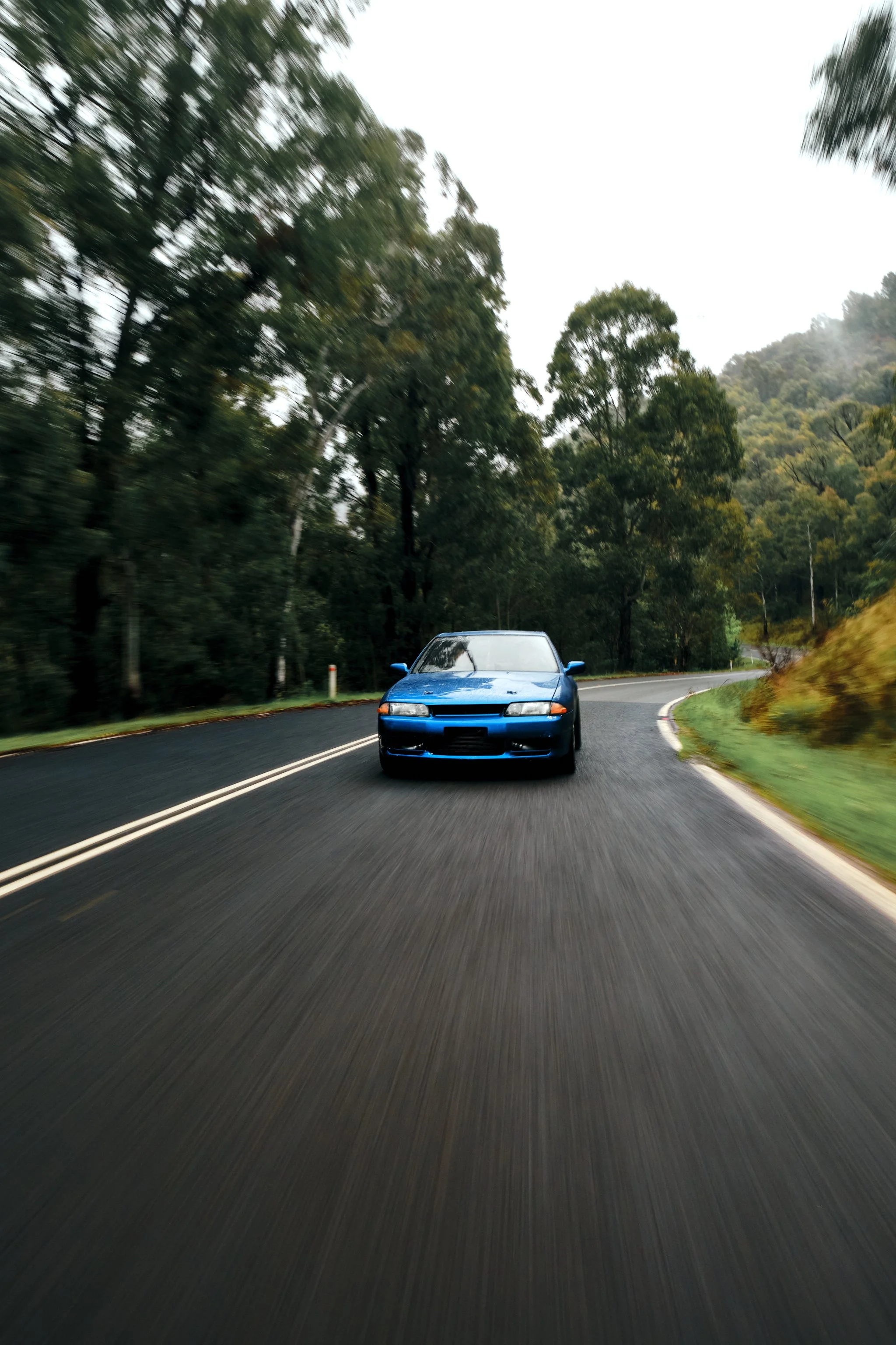 A blue sports car driving on a curvy mountain road surrounded by trees and greenery on a cloudy day.