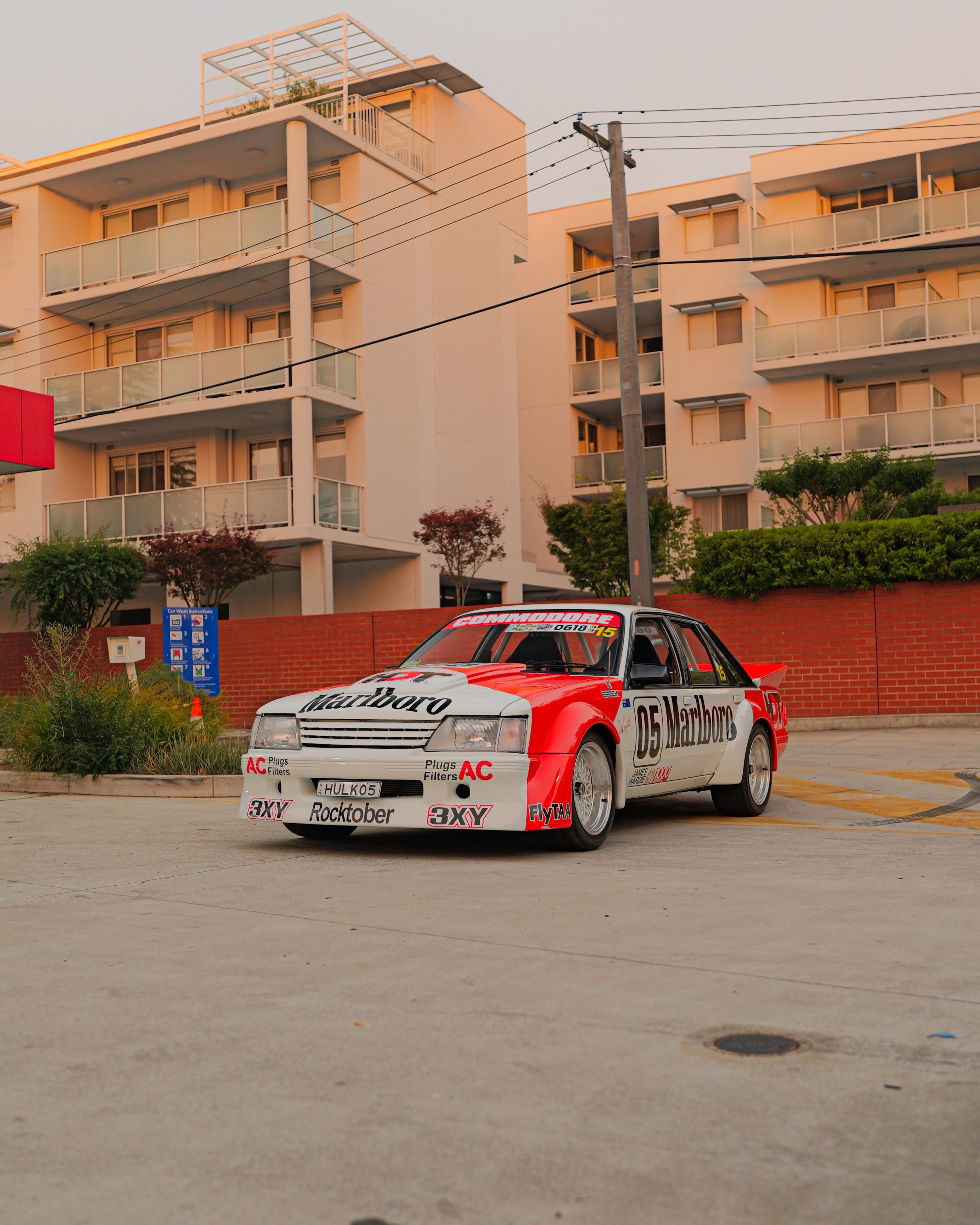 A red and white racing car with sponsor logos parked in a parking lot in front of an apartment building with trees and a blue car wash sign nearby.