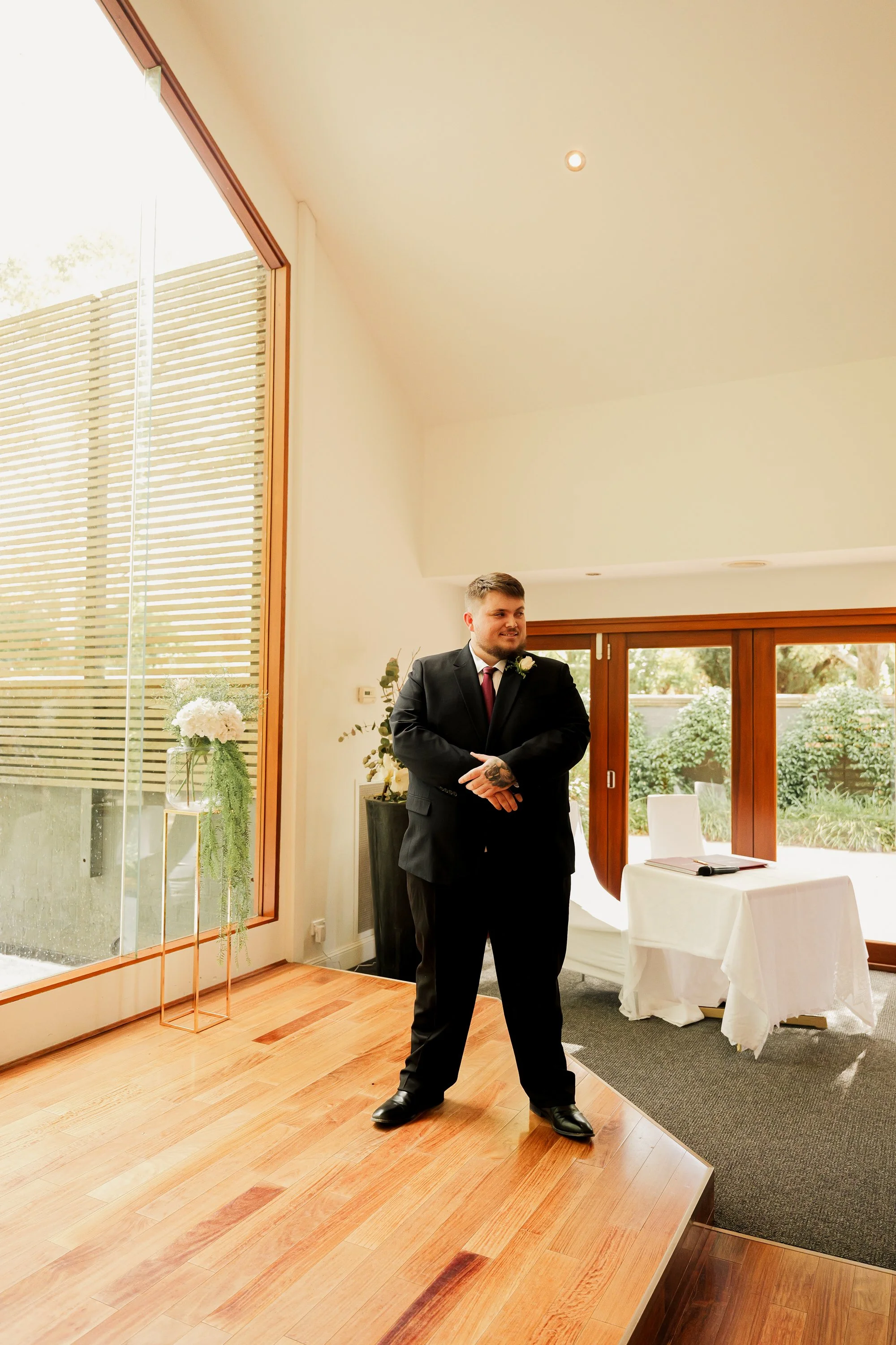 A man in a suit and tie standing in a room with large windows, next to a table with a notebook, during a wedding or formal event.