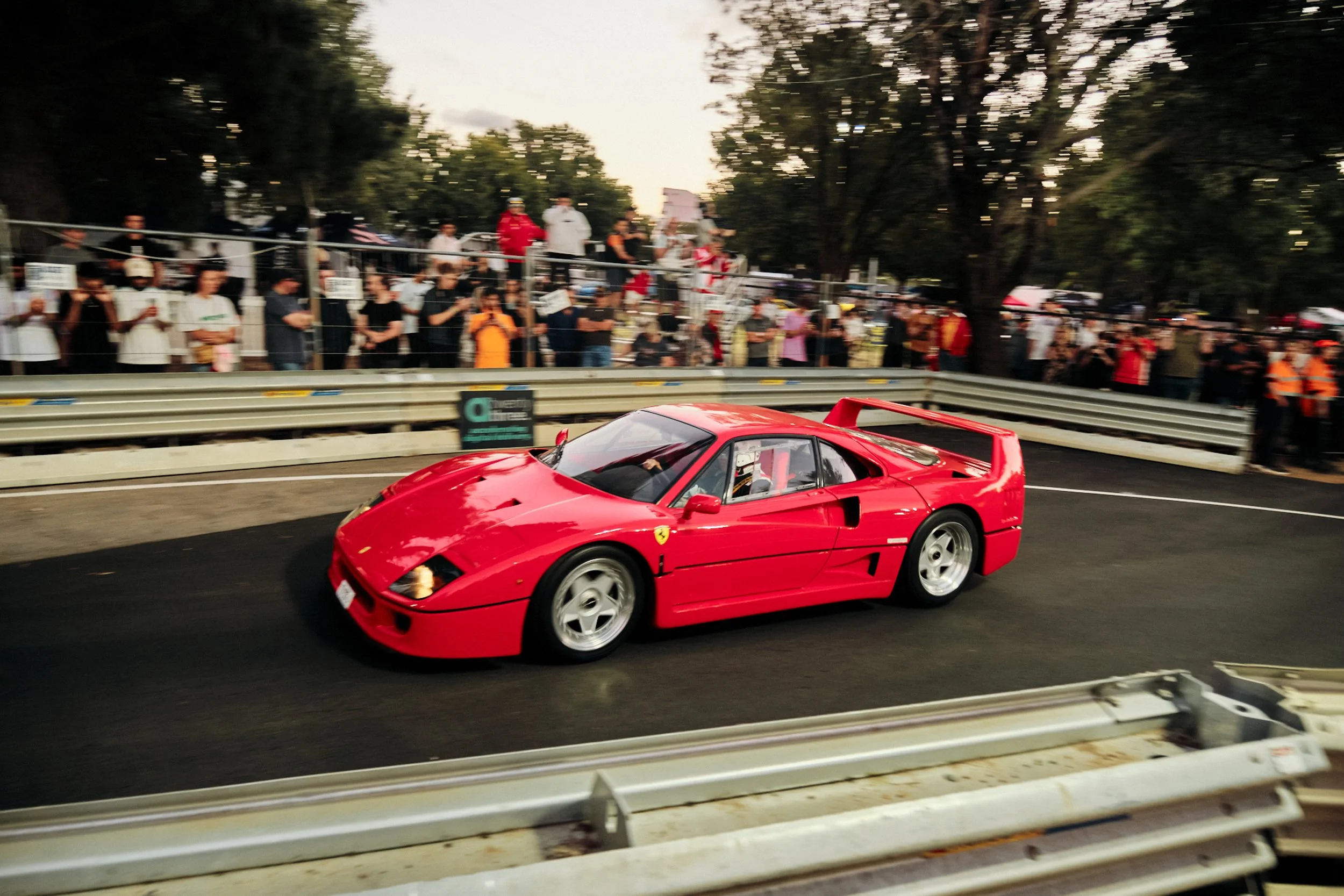 A red Ferrari sports car racing on a track with a crowd of spectators watching from the sidelines behind a fence.