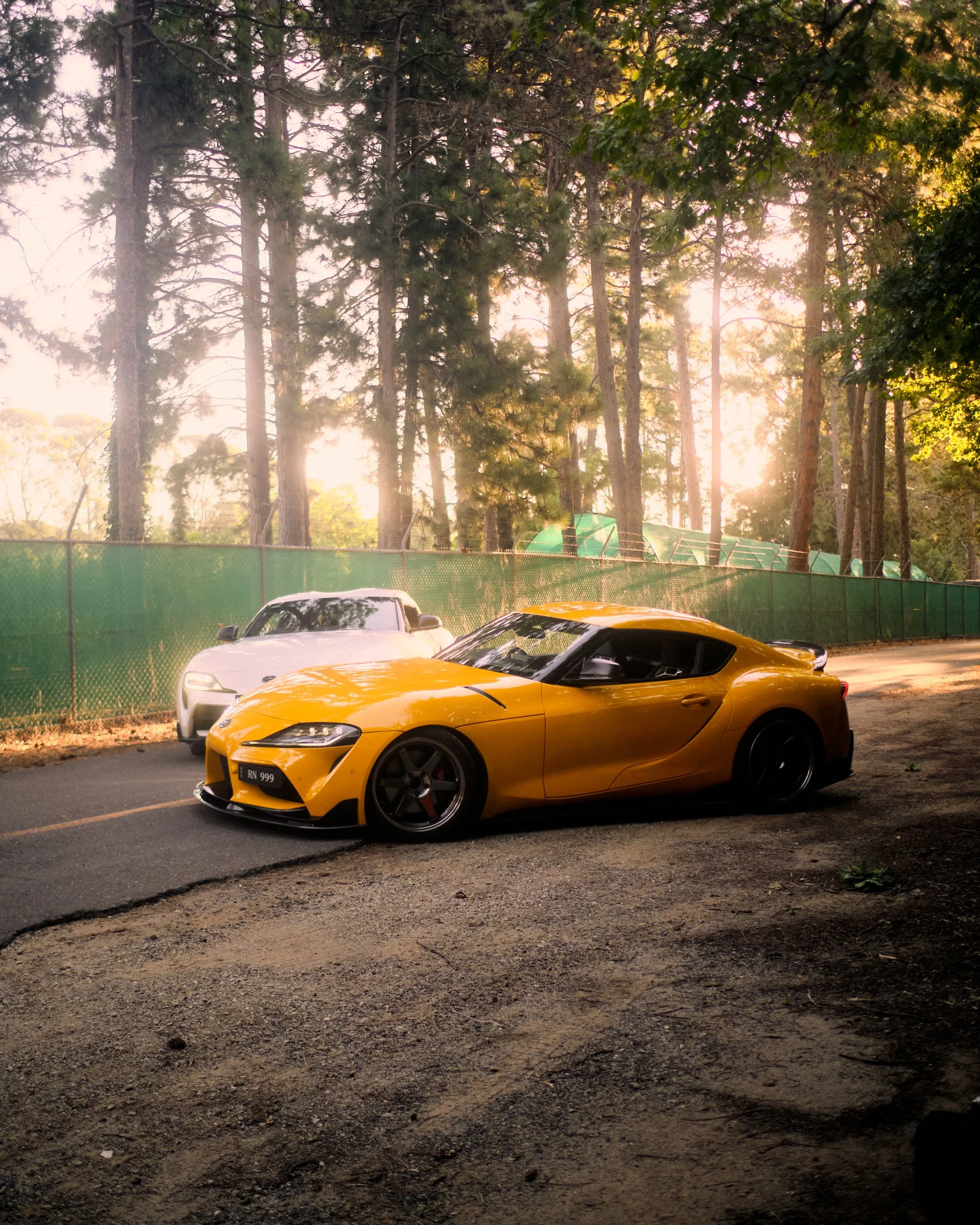 A bright yellow sports car parked on a dirt lot with a black roof and red brake calipers, adjacent to a silver car and behind a chain-link fence. Tall trees with sunlight filtering through the leaves are in the background.