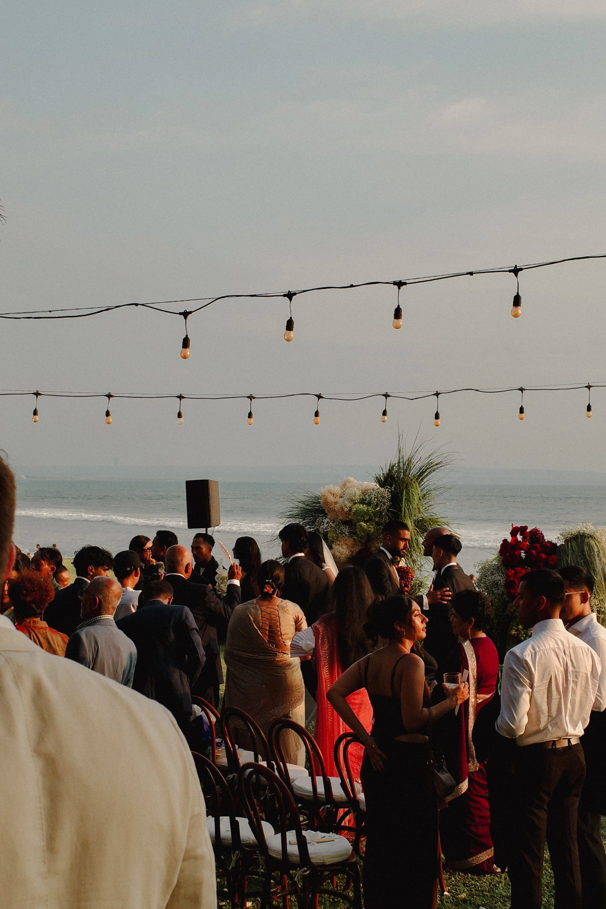 People gathered outdoors for a wedding or celebration near the ocean, with string lights hanging in the sky and flowers in the background.