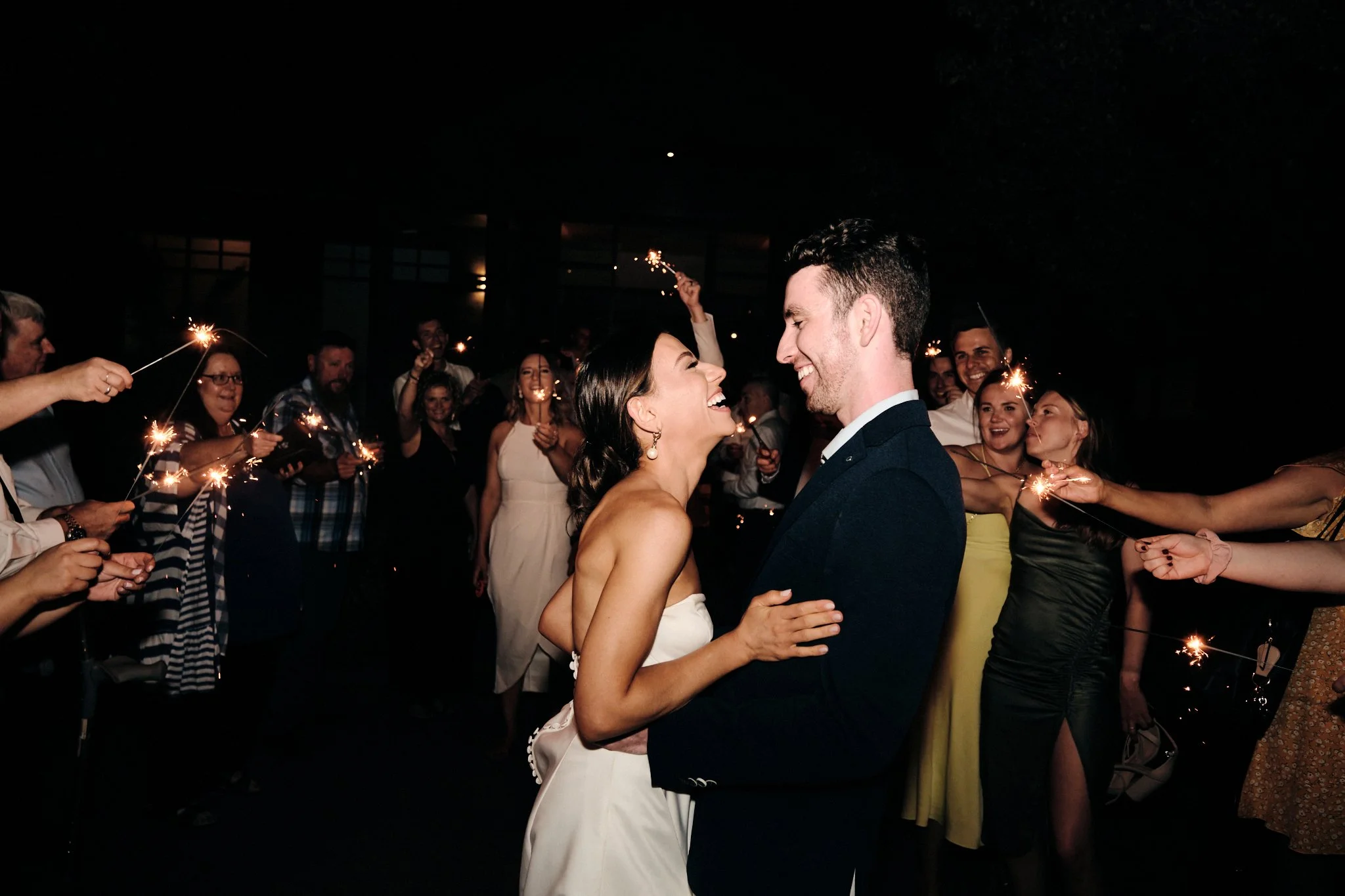 A bride and groom smiling and looking into each other's eyes during their wedding celebration, surrounded by guests holding sparklers at night.