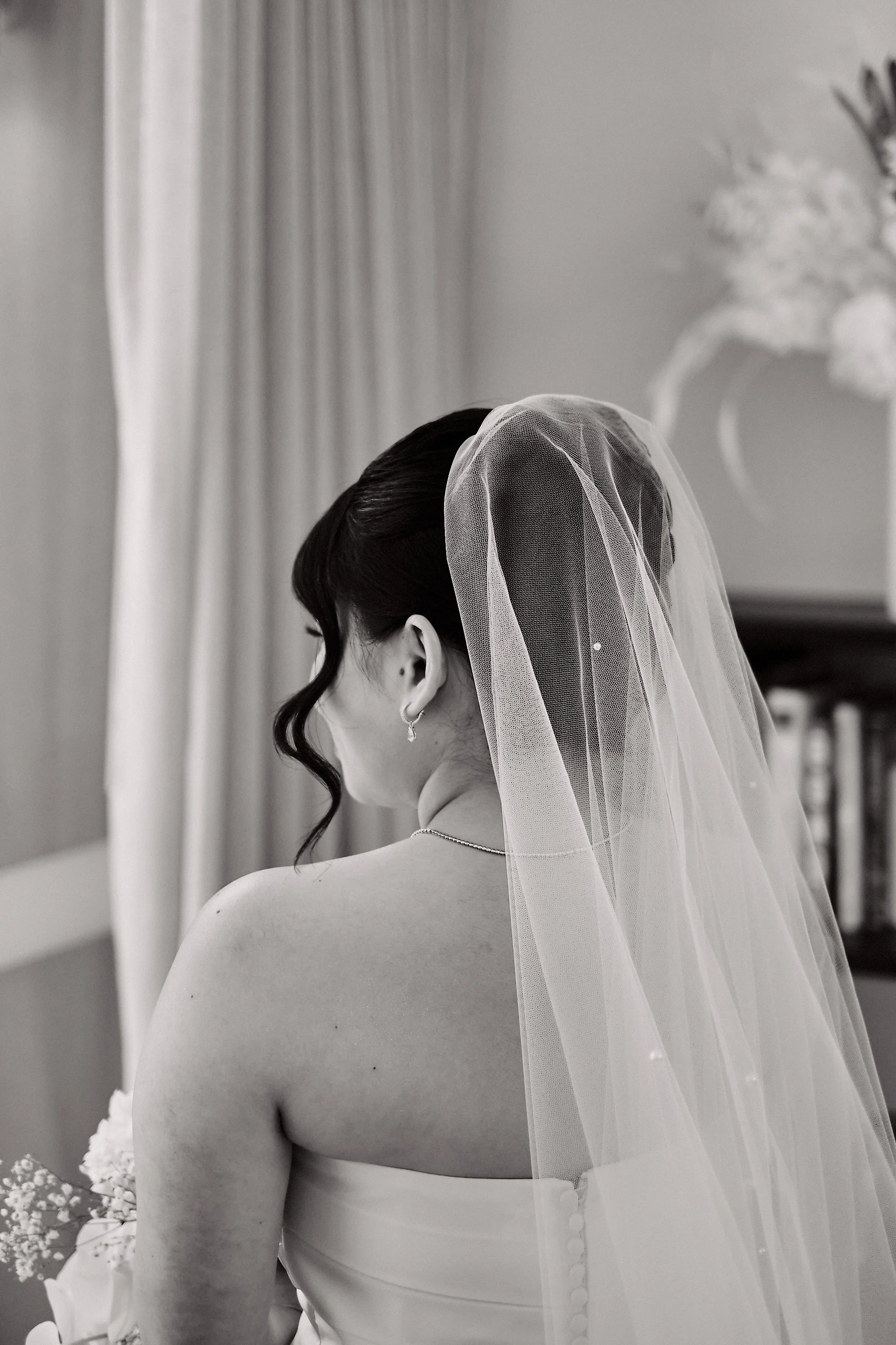 Black and white photo of a bride with dark hair styled in an updo with a veil, wearing earrings, and a strapless dress. She is posed with her back to the camera, showing the back of her dress and veil. There are flowers visible on the left side and a