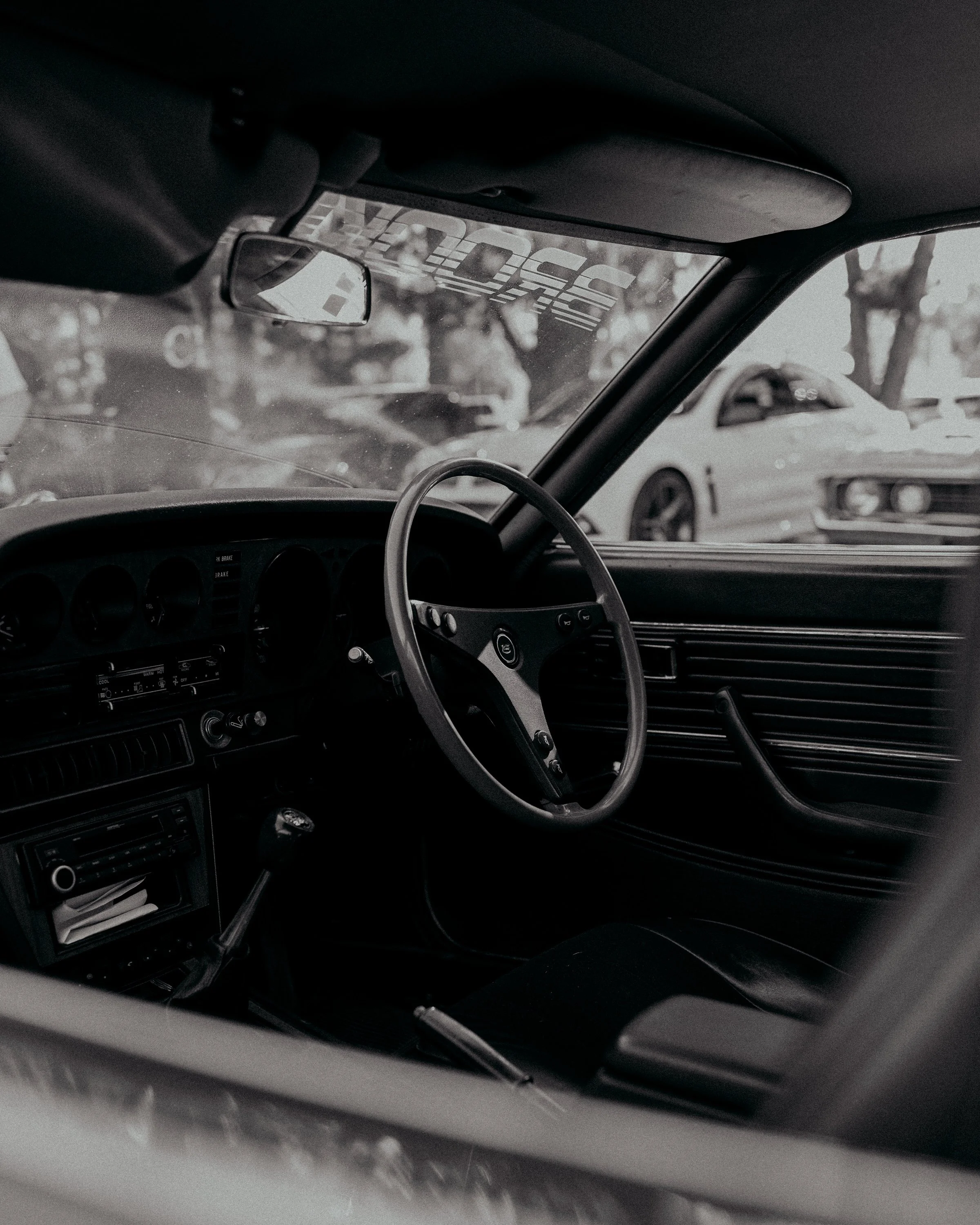 The photo shows the interior of a vintage car, focusing on the dashboard, steering wheel, and rearview mirror. Through the windshield, other cars and trees are visible in a parking lot or outdoor setting.