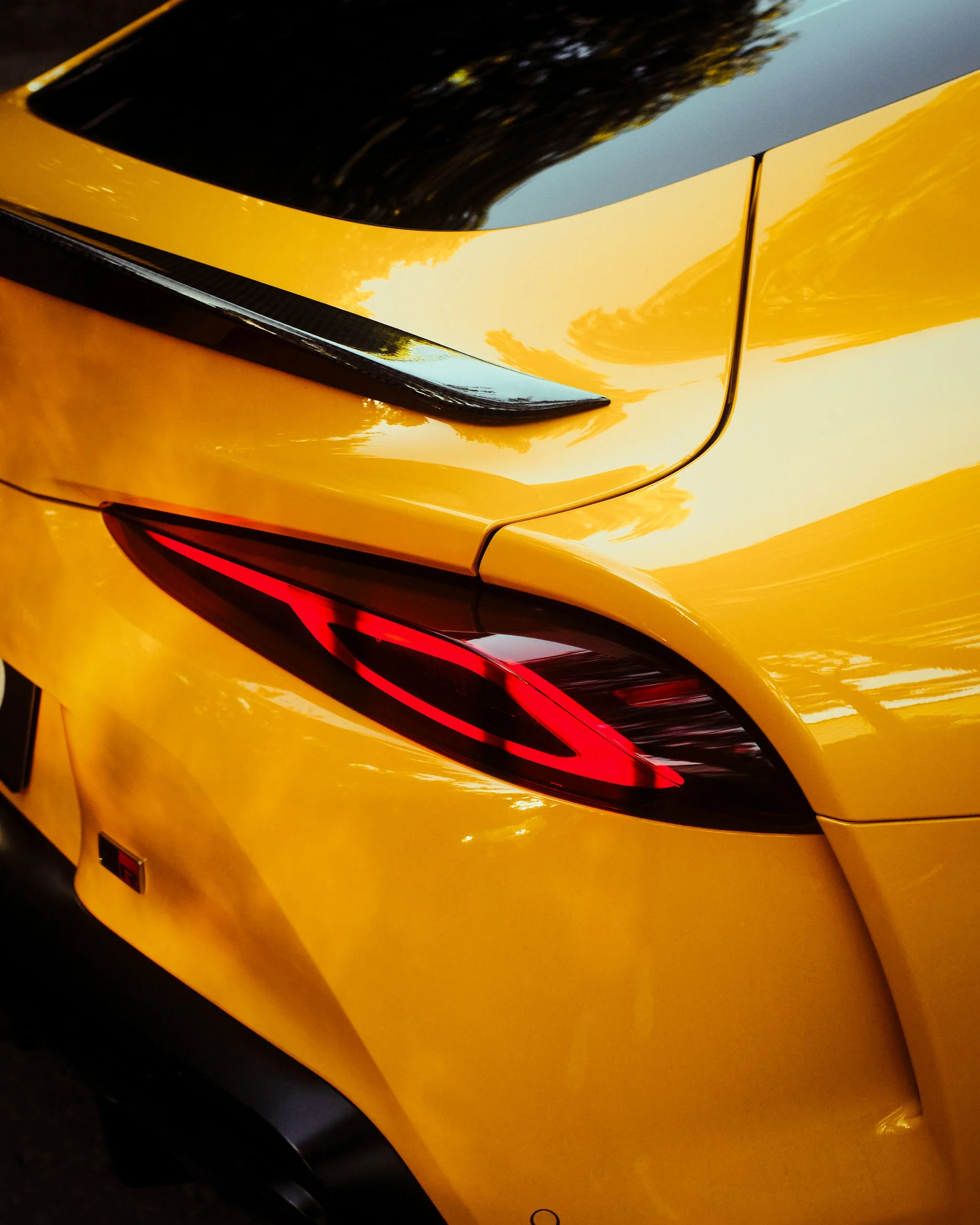 Close-up of the rear end of a yellow sports car, showing part of the taillight and rear quarter panel with reflections of trees and sky.