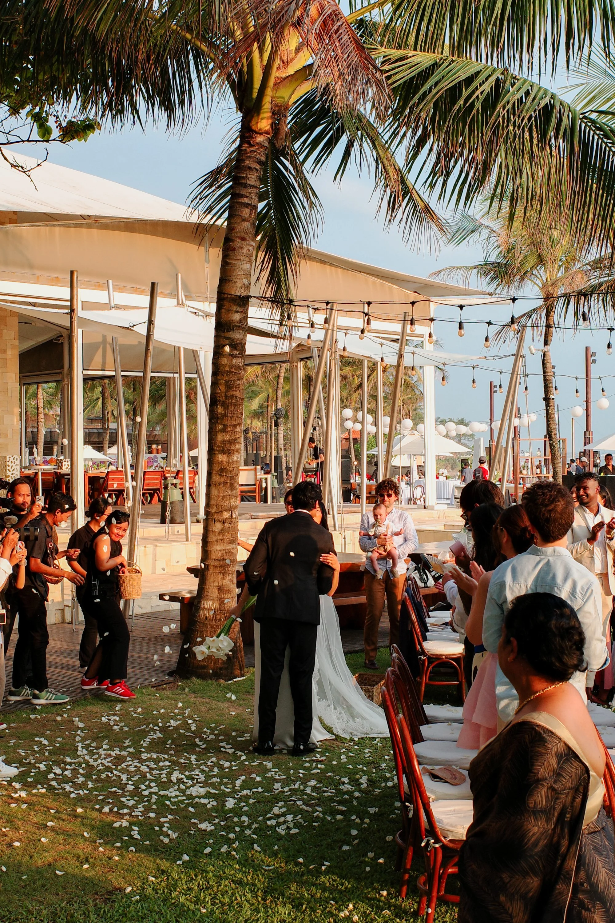 A wedding ceremony outdoors with guests seated and standing, palm trees, white flower petals on the ground, and string lights hanging in the background during daytime.