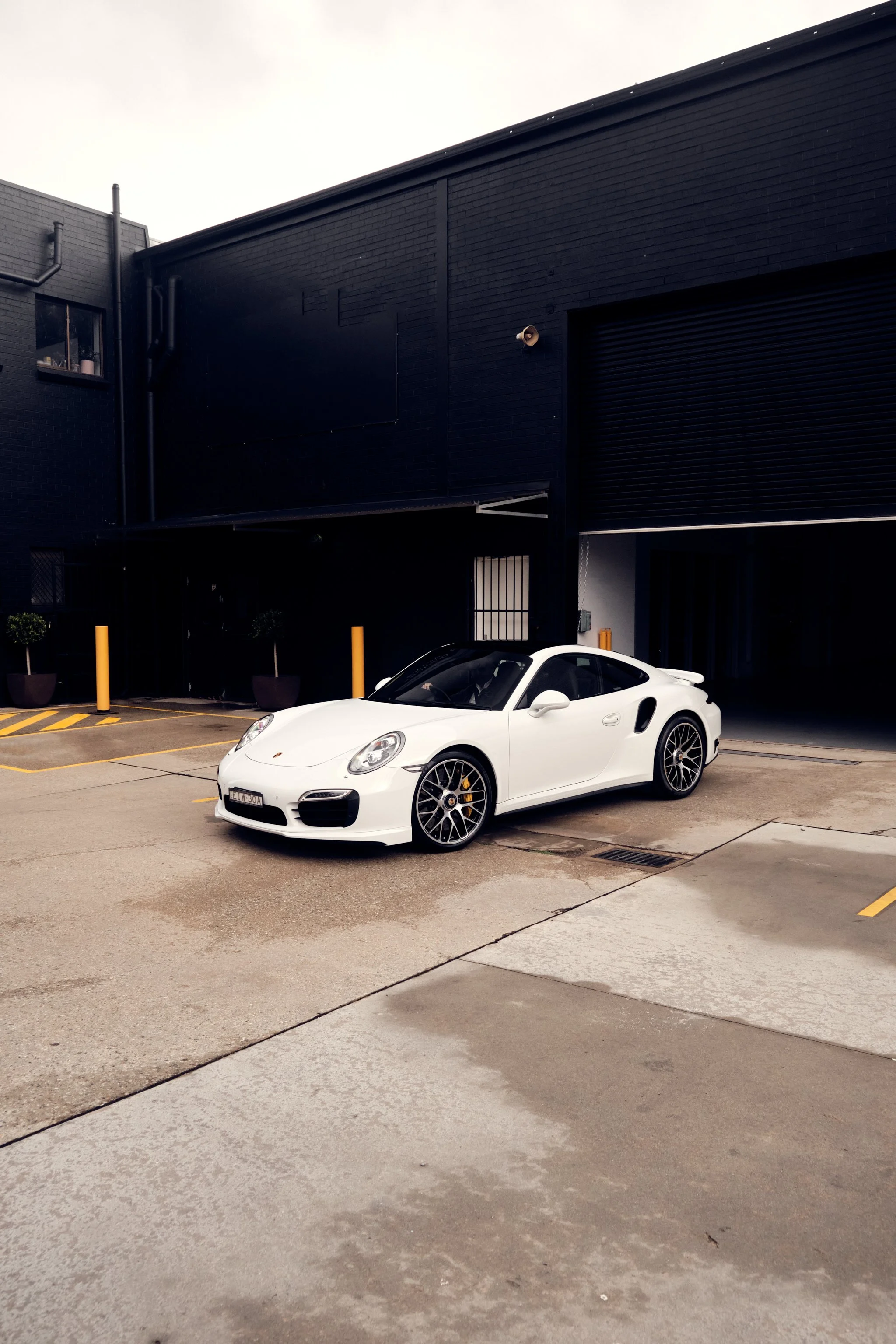 A white sports car parked in a parking lot in front of a black building with a black roller door and black walls. The sky is overcast.