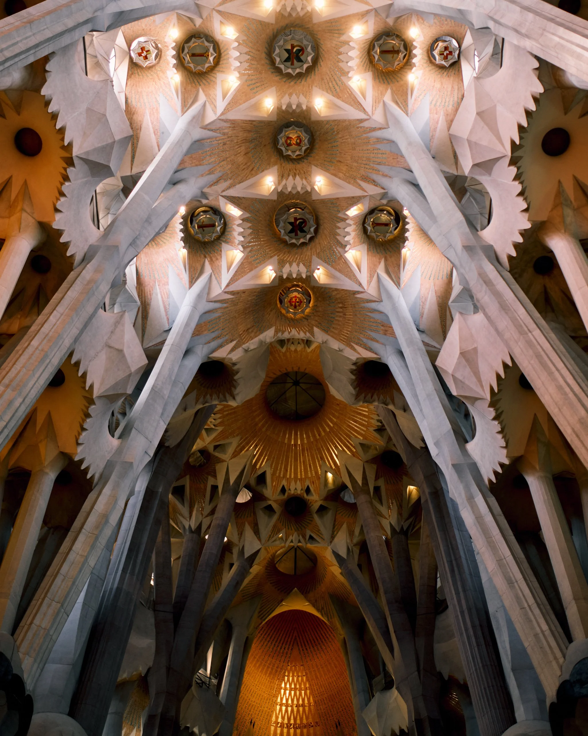 The ceiling of Sagrada Familia basilica with intricate geometrical patterns, illuminated by small lights, and featuring religious symbols at the center of each decorative element.