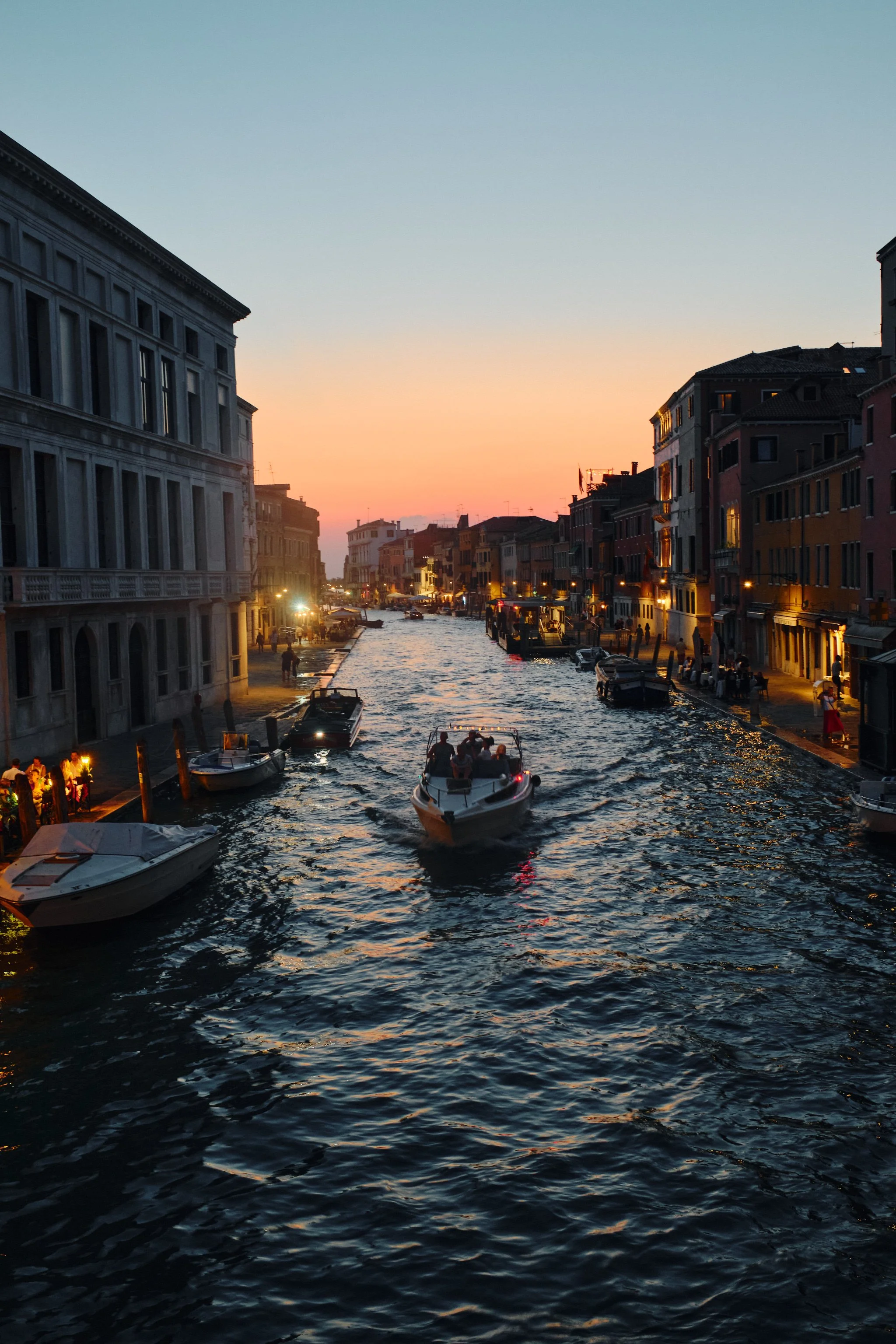 A canal in Venice at sunset with boats on the water, historic buildings on both sides, and people walking along the promenade.