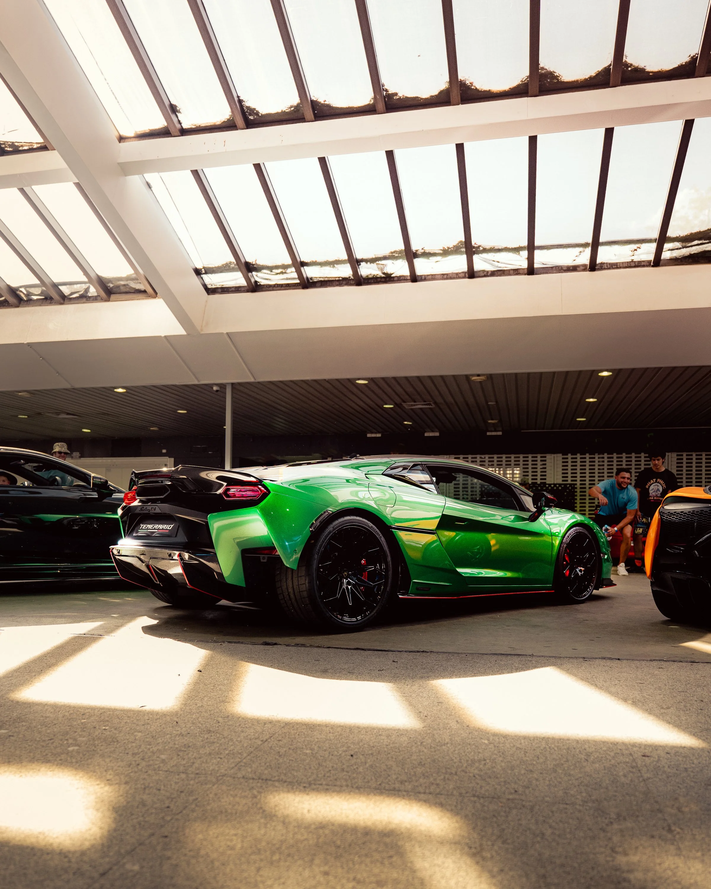 A bright green sports car with black rims and racing stripes parked indoors, with a large skylight overhead and people around.