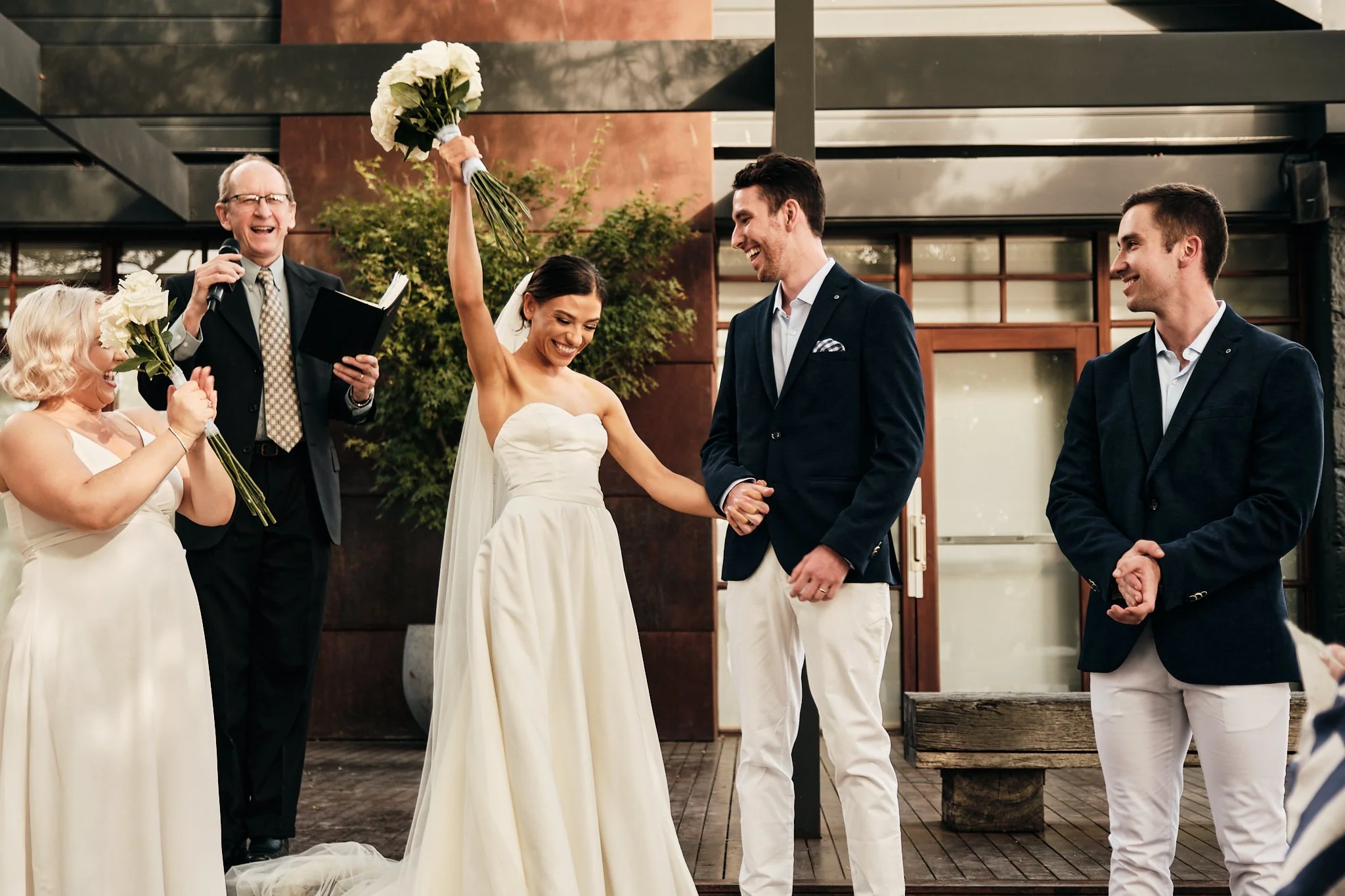 A wedding ceremony where a woman in a white dress is holding a bouquet of white flowers in the air, smiling, while a man in a navy blazer and white pants is holding her hands and also smiling. Two women in white dresses hold bouquets, and a man in a 