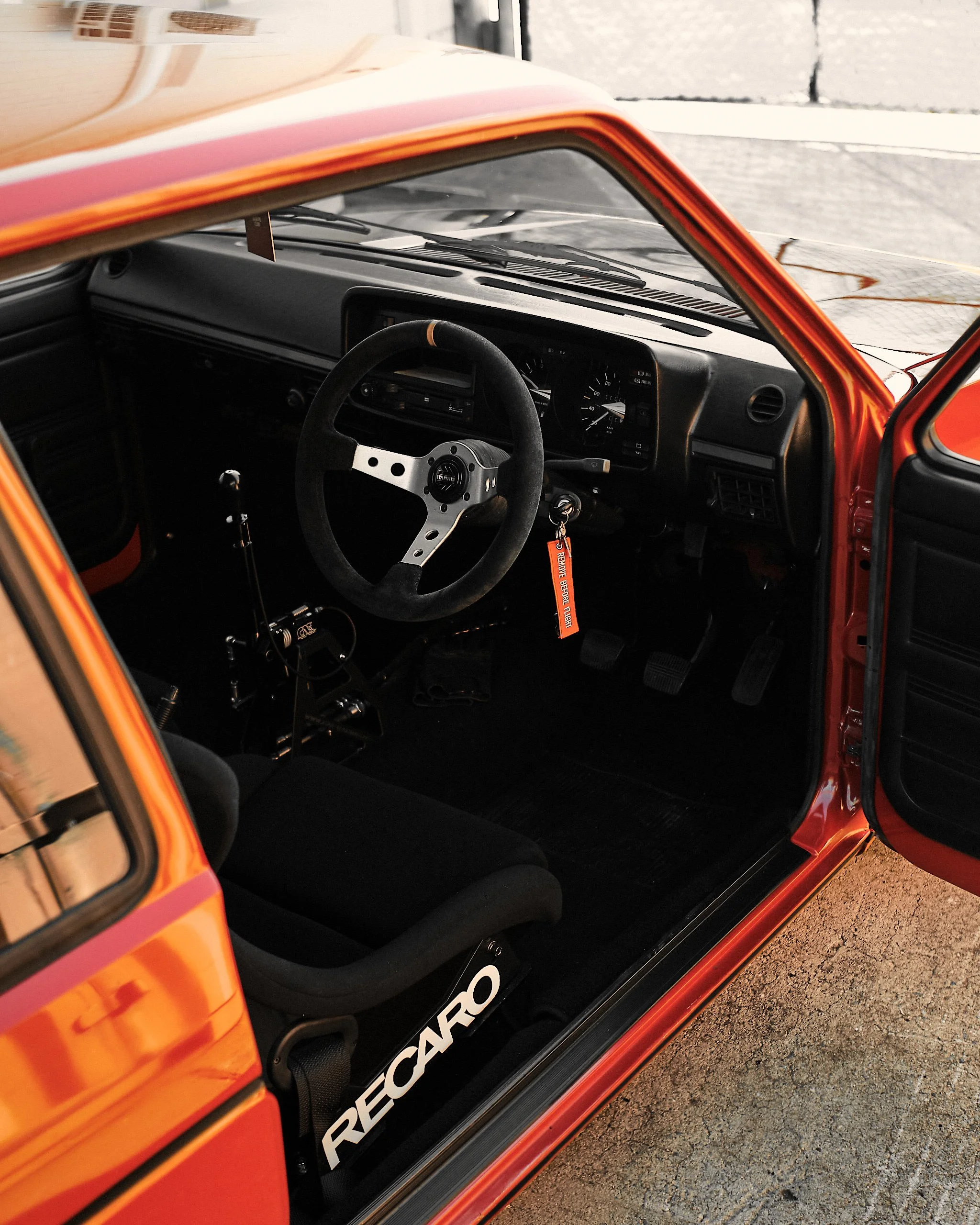 Interior of a race car with a racing steering wheel, dashboard, pedals, and a black seat, viewed from the passenger side with part of the red exterior visible.