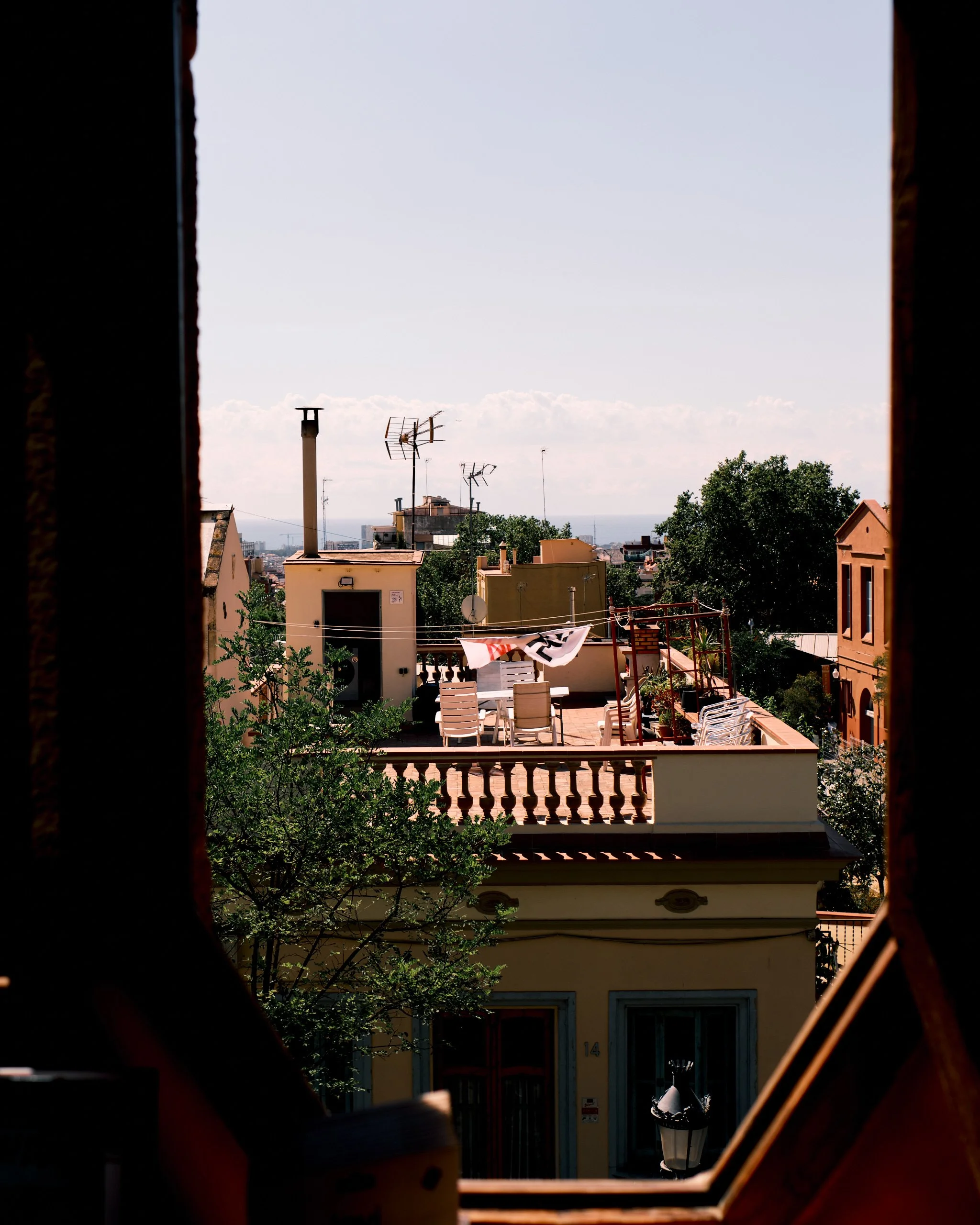 View through a window opening showing rooftops, television antennas, chimneys, trees, and a bright sky in an urban area