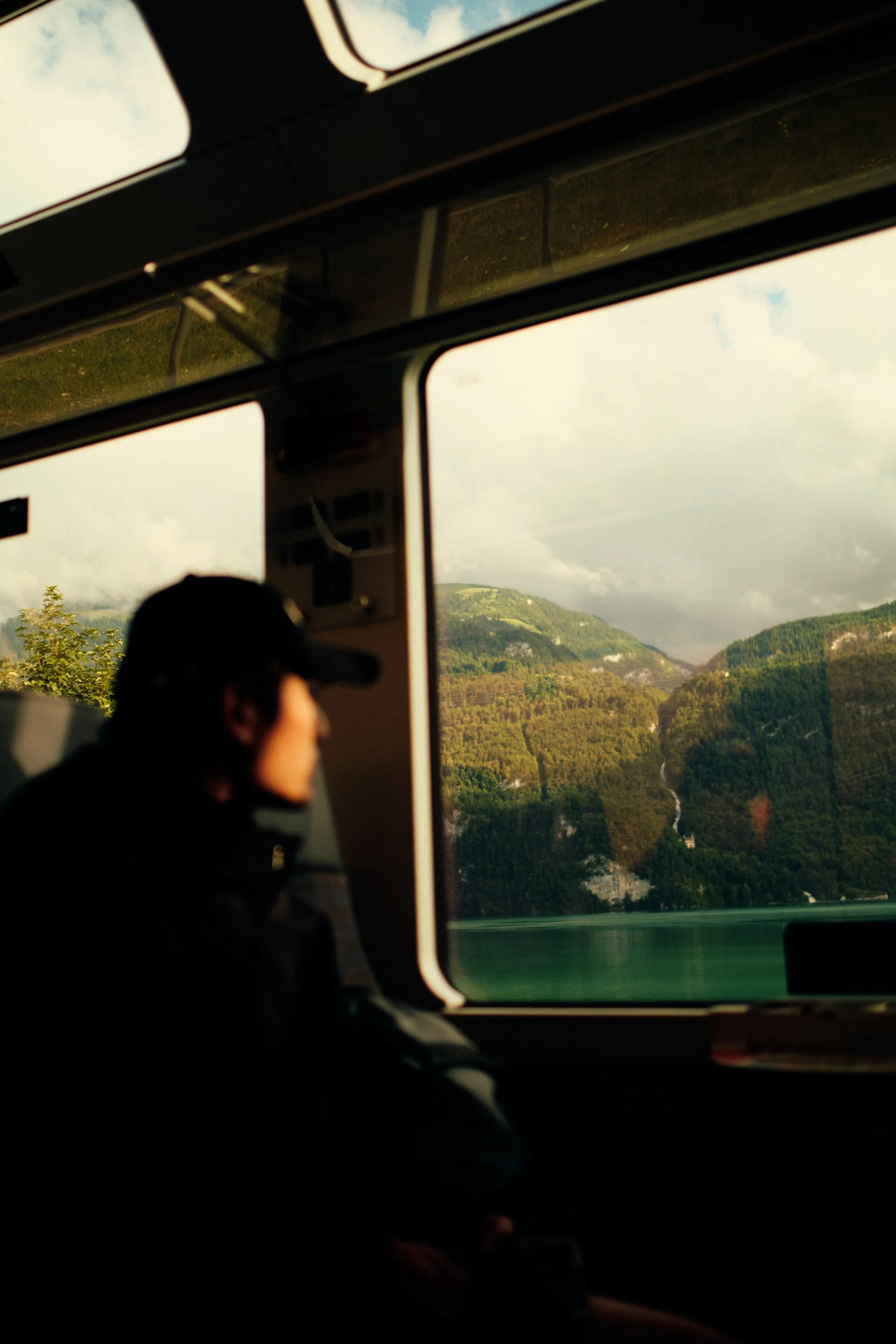 Photo taken inside a bus showing a reflection of a person wearing a dark cap, overlooking a landscape of green hills, a river, and cloudy skies through the window.