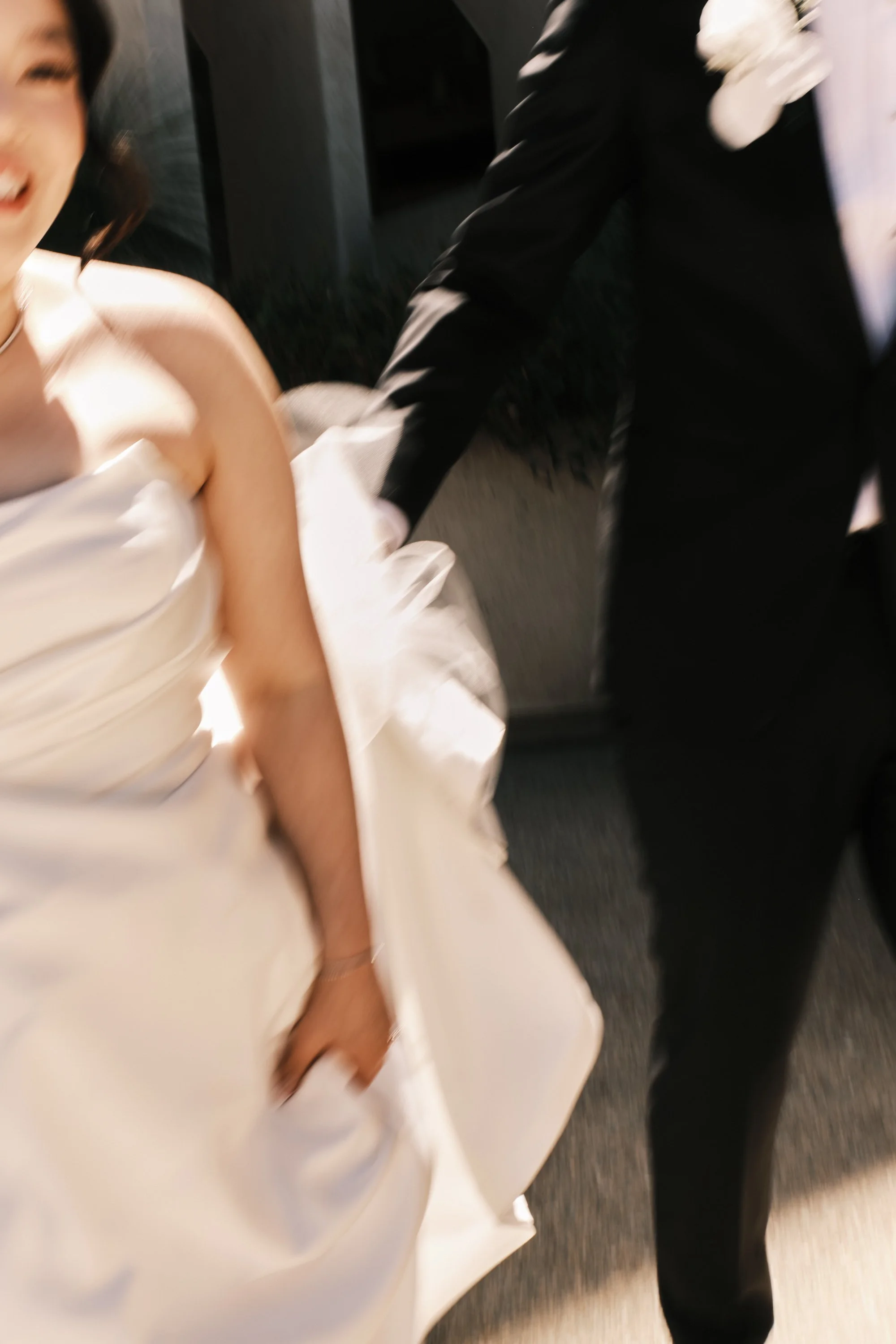 A young woman in a strapless white wedding dress with a satin finish, sitting and smiling, with a person in formal attire (possibly a tuxedo) holding her hand.