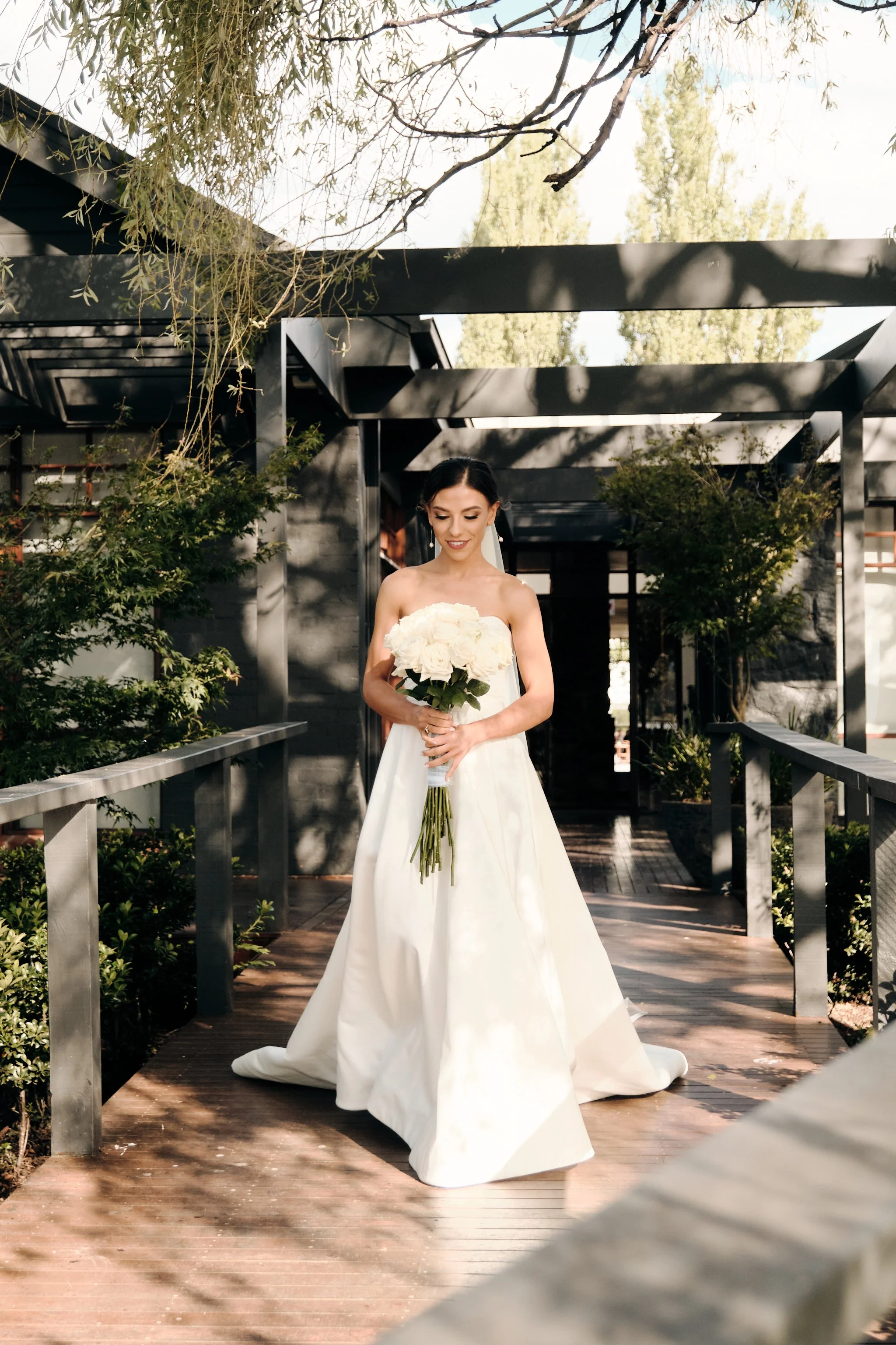 A bride in a white wedding dress holding a bouquet of white roses, standing on a wooden pathway with greenery and modern black structures around her.