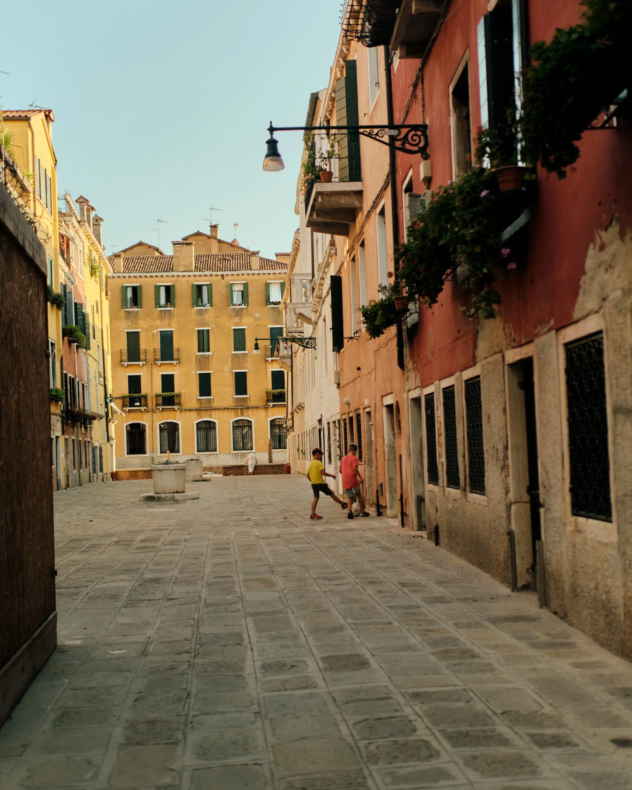 A narrow alleyway in an Italian city with colorful buildings, flower pots on balconies, hanging street lamps, and a few children playing in the distance on a sunny day.