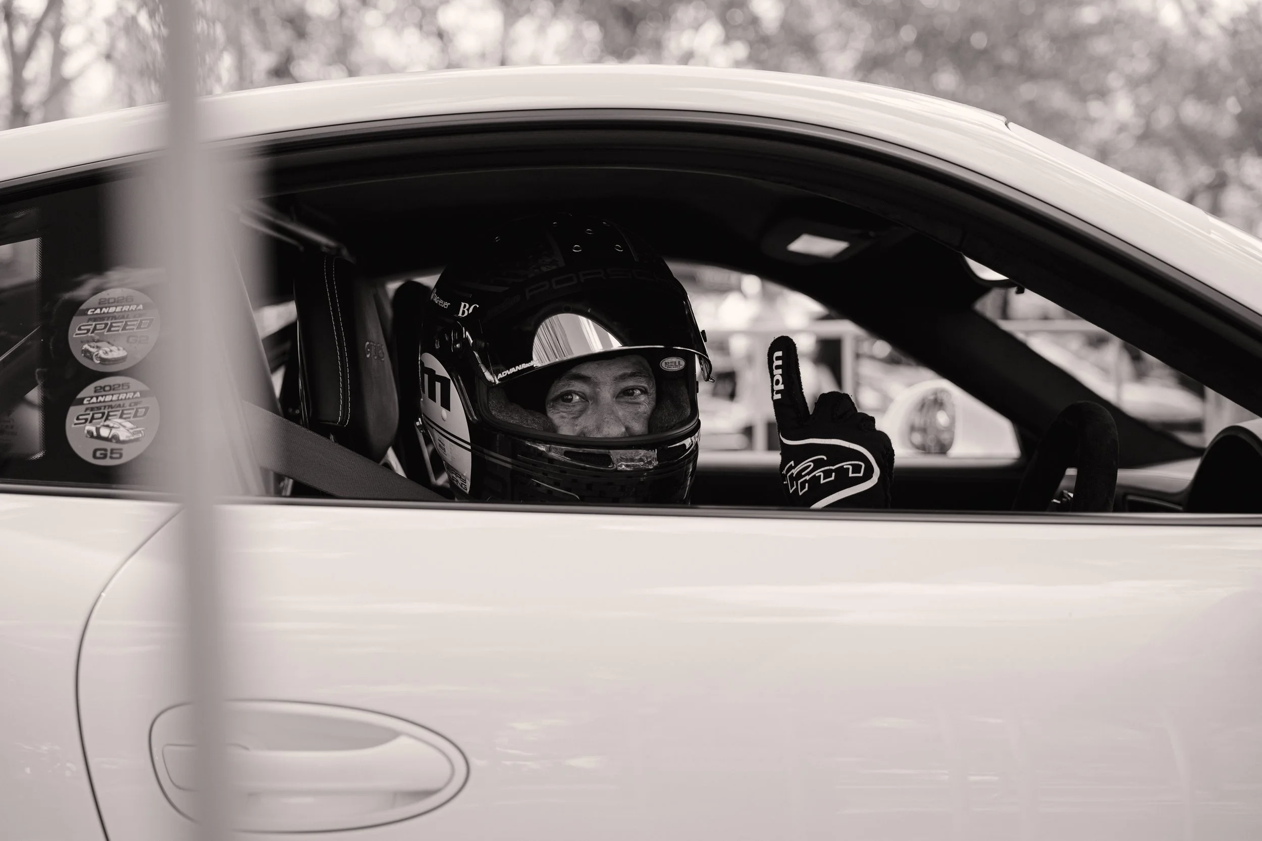 A race car driver sitting inside a race car, wearing a helmet and racing suit, giving a thumbs-up gesture. The car's interior and part of the exterior body are visible.