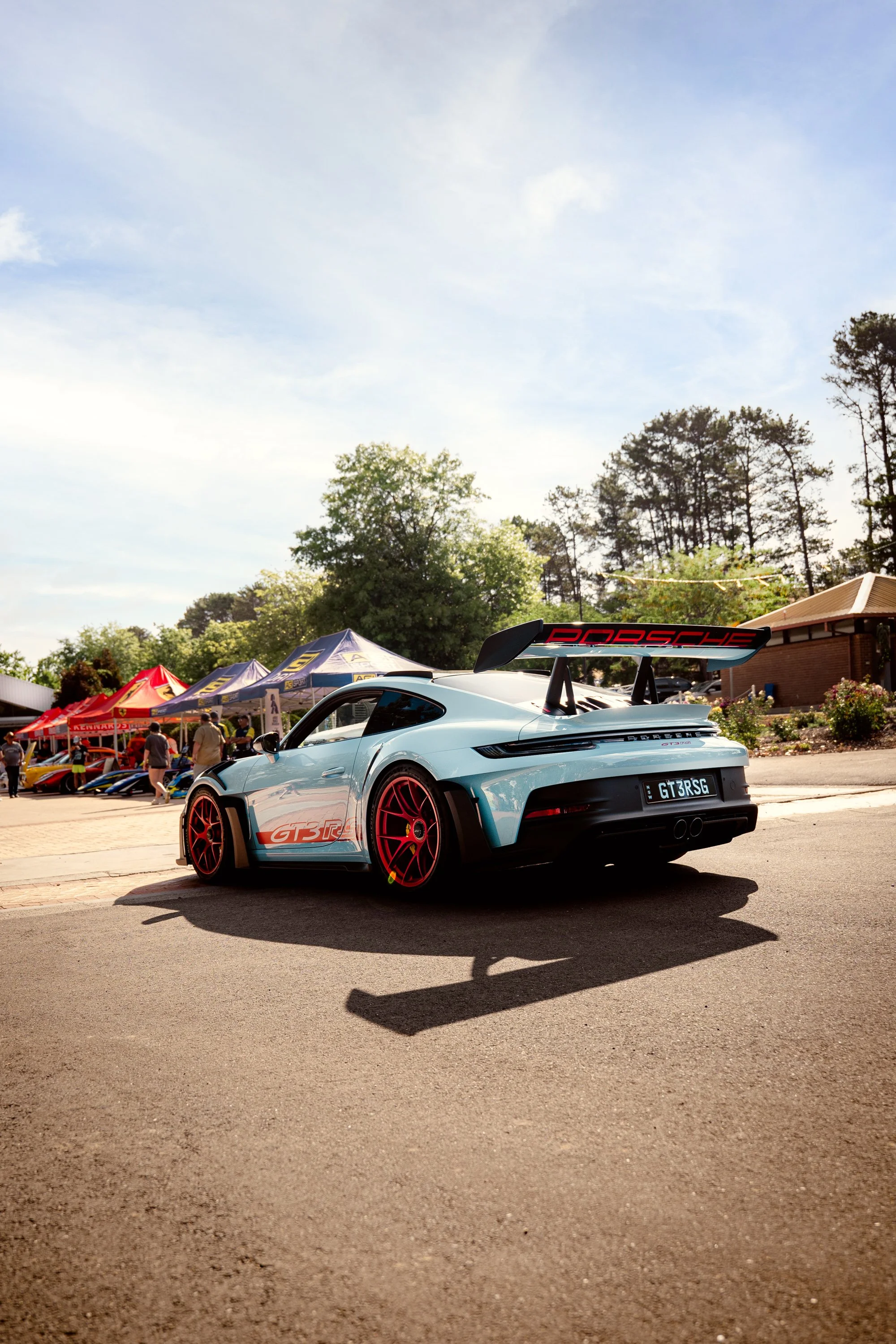 A light blue Porsche 911 GT3 RS with red wheels and a large rear wing, parked outdoors at a car show. Tents and people are visible in the background, along with trees and a building under a partly cloudy sky.