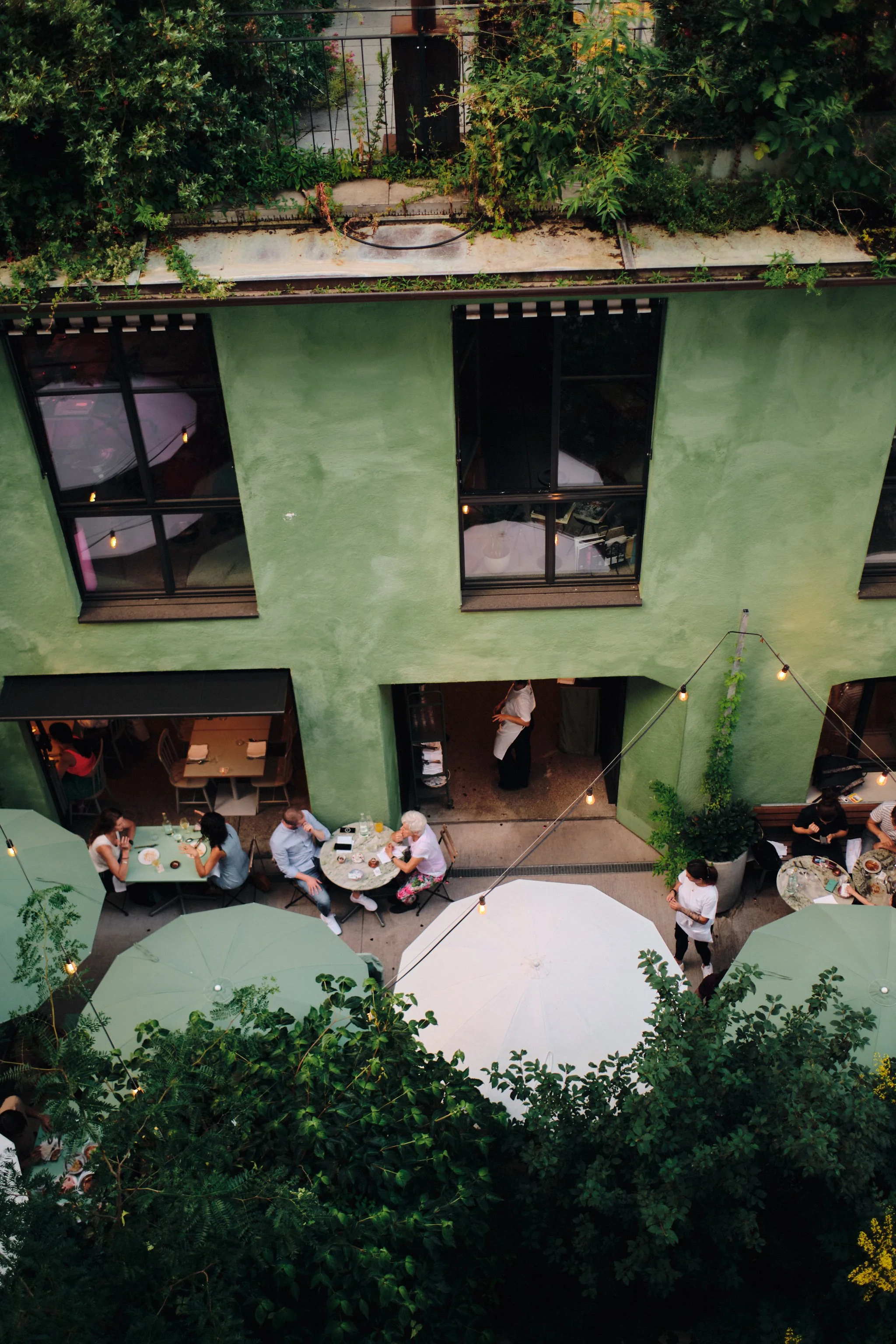 Outdoor dining scene with people sitting at tables with white and green umbrellas, greenery around, and a building with green walls and large windows.
