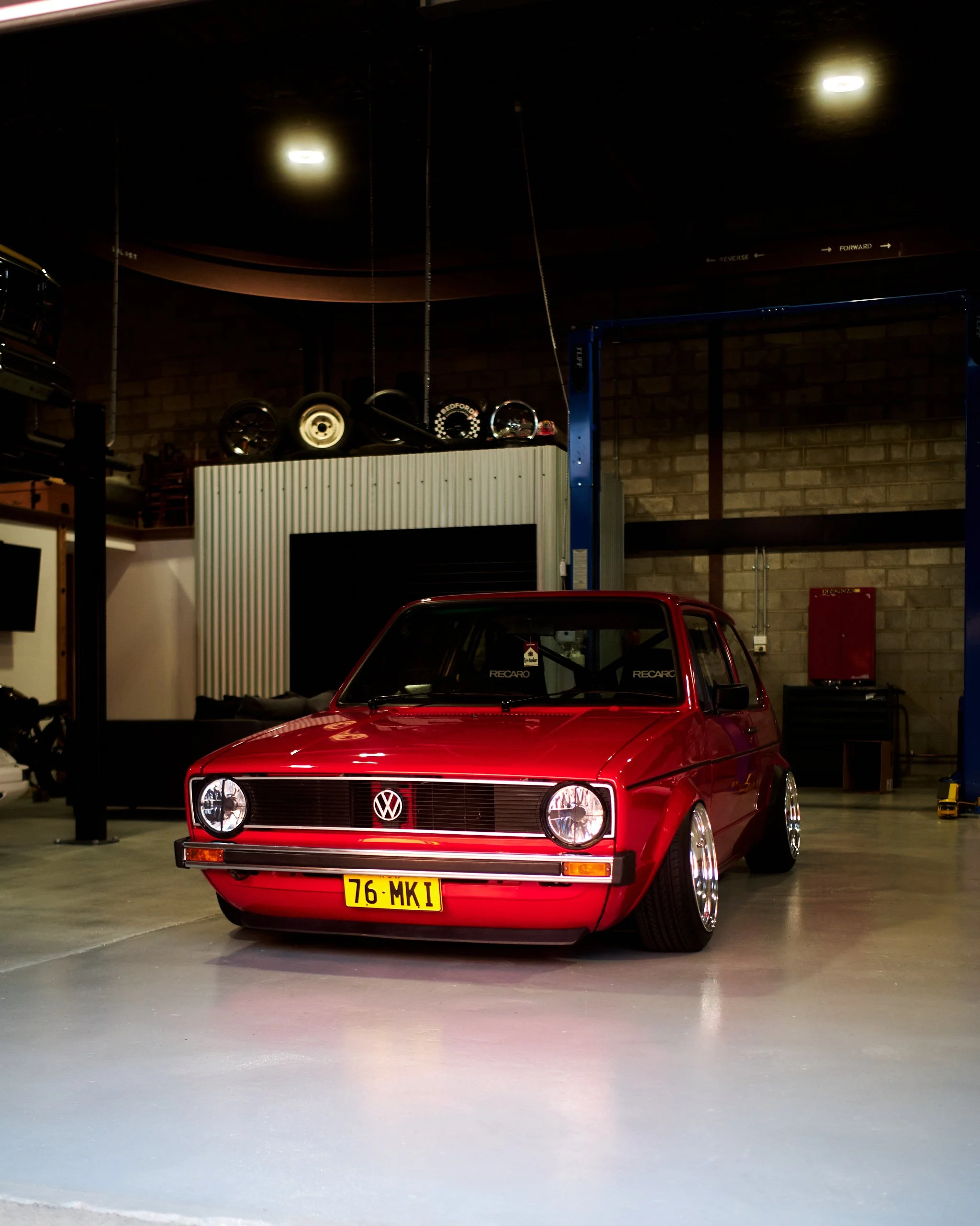 Red vintage Volkswagen Golf parked inside a spacious garage or warehouse with a polished concrete floor, brick and metal walls, and industrial lighting. A car lift is partially visible on the right, and tires and automotive parts are stored on a shel