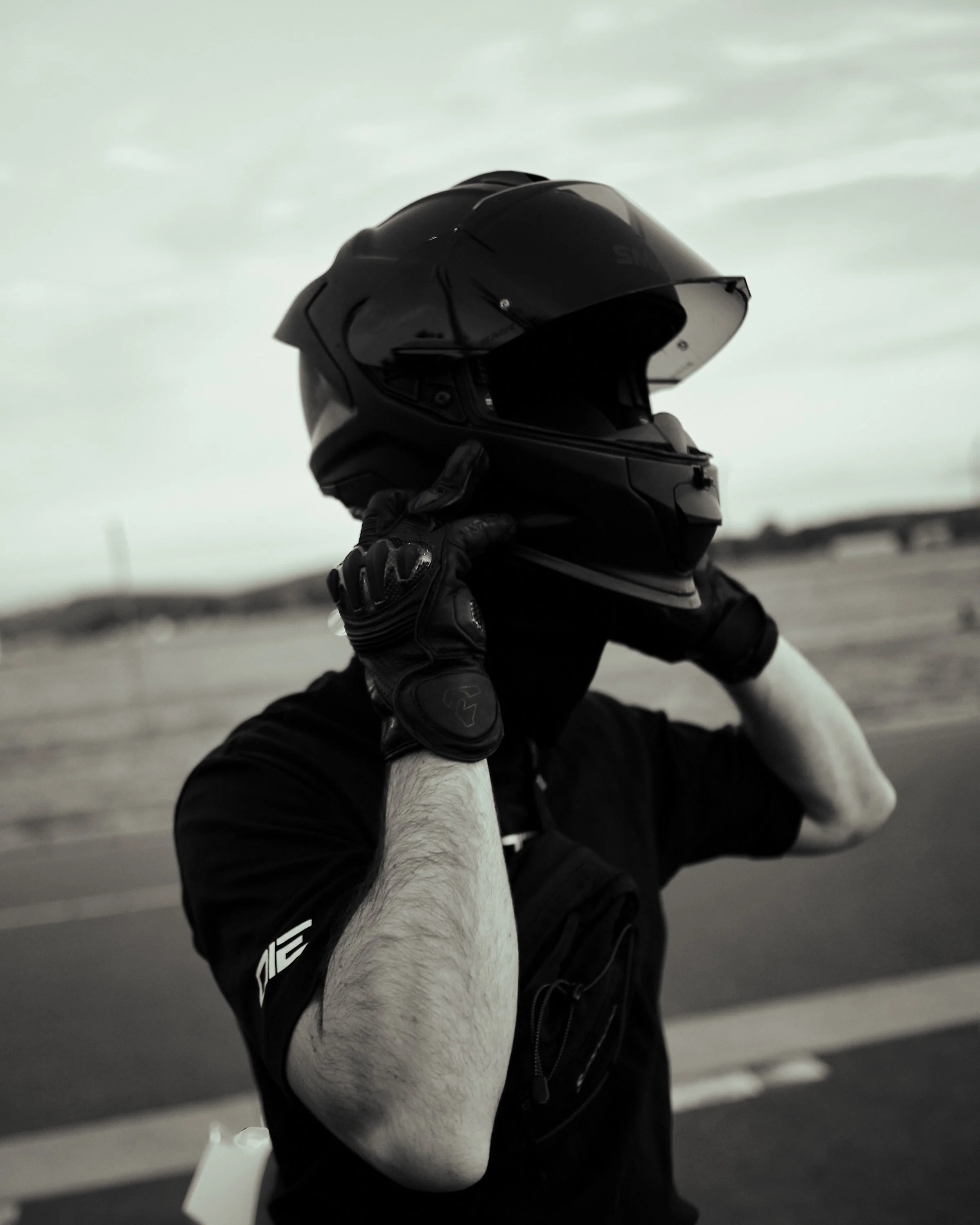 Person holding a motorcycle helmet with one hand, wearing a black jacket and gloves, with a dog lying on the ground, on a road with a cloudy sky in the background.