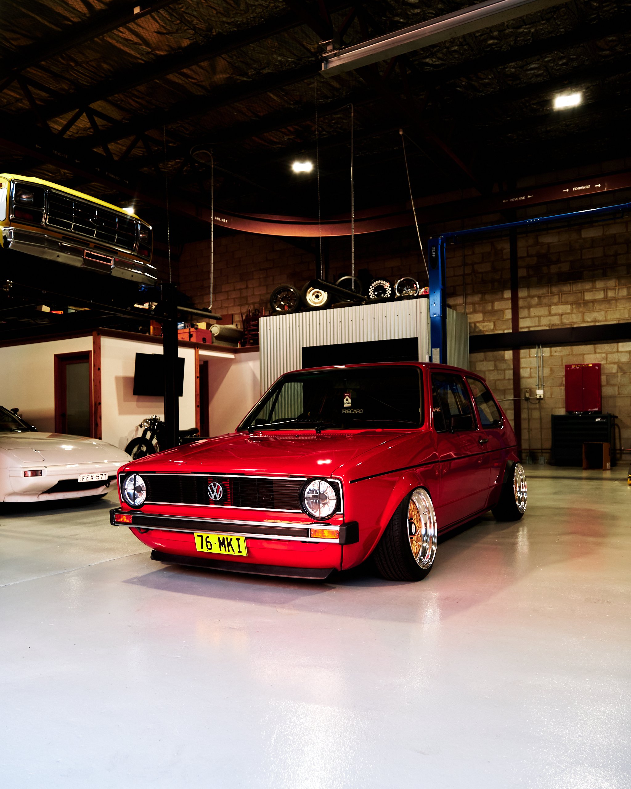 Red vintage Volkswagen car in a garage with other classic cars and car wheels hanging on the wall.
