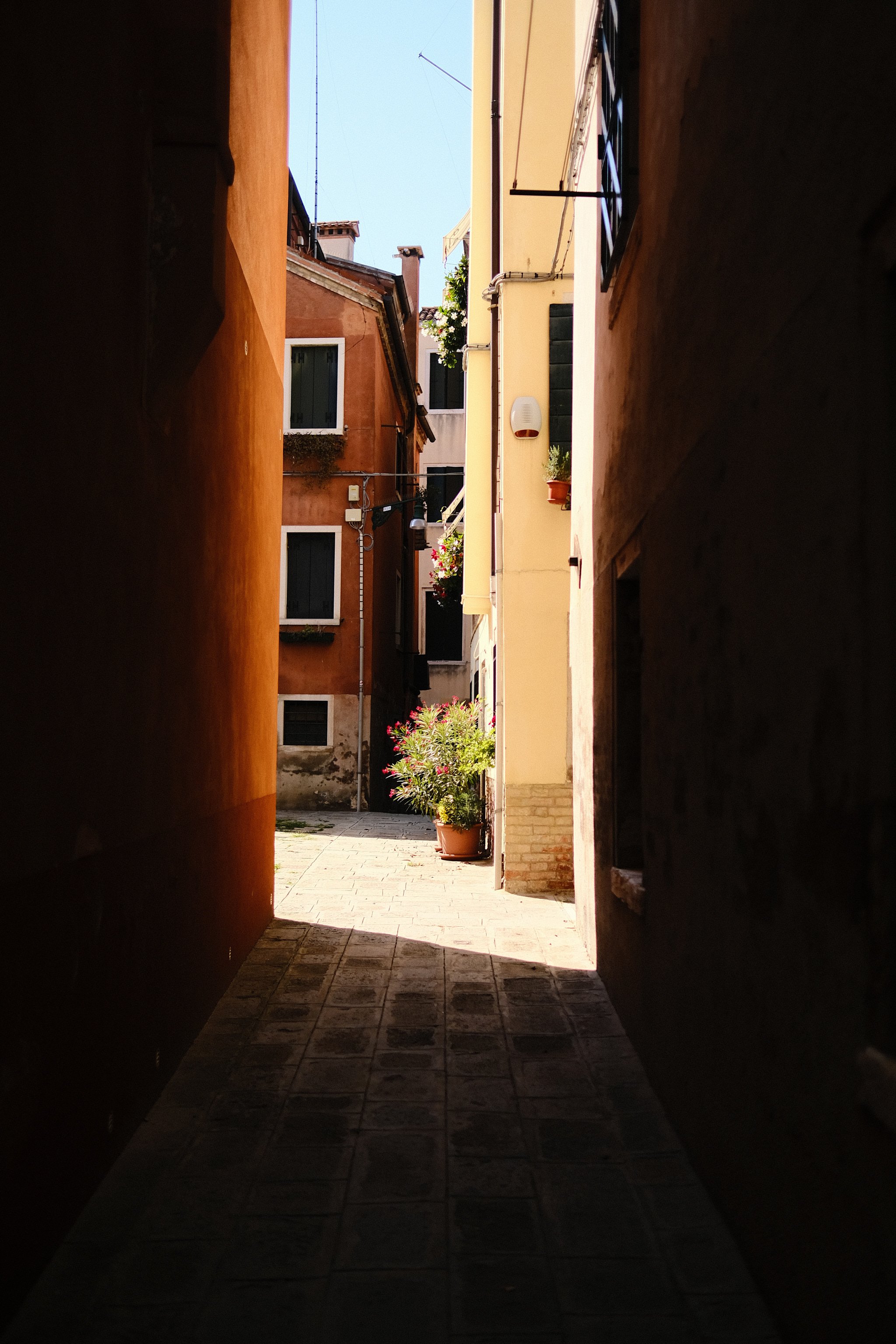 A narrow alleyway between buildings leads to a sunlit courtyard with potted plants and colorful flowers.