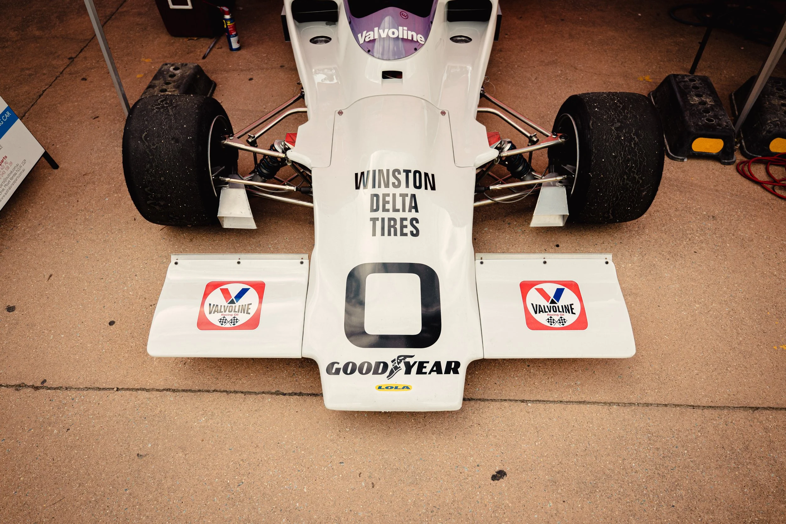 A vintage Formula 1 race car displayed on a concrete surface. The car is mostly white with various sponsor logos, including Valvoline, Goodyear, and Lola. The front view shows large black tires, suspension components, and a canopy with the text 'Wins