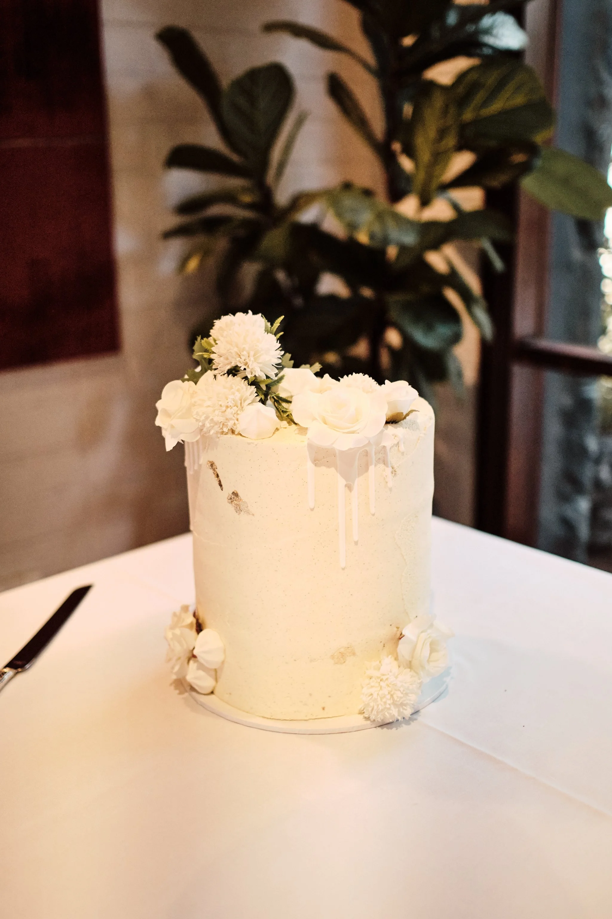 A tall, white, cylindrical cake decorated with white flowers and floral accents, sitting on a white table with a cake knife nearby. In the background, a large green plant is seen indoors.