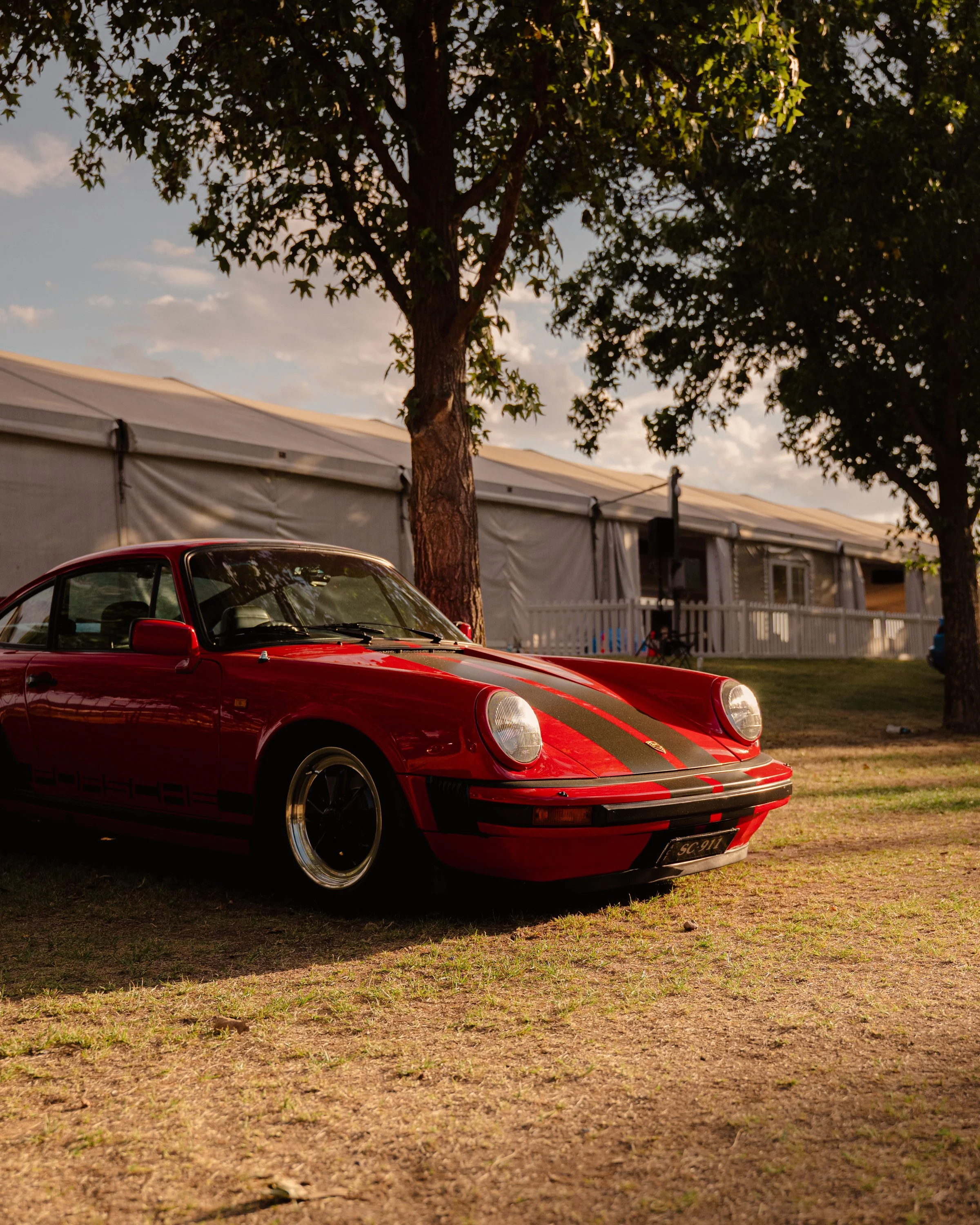 A red sports car with black racing stripes parked on grassy ground near trees and a large event tent under a partly cloudy sky.