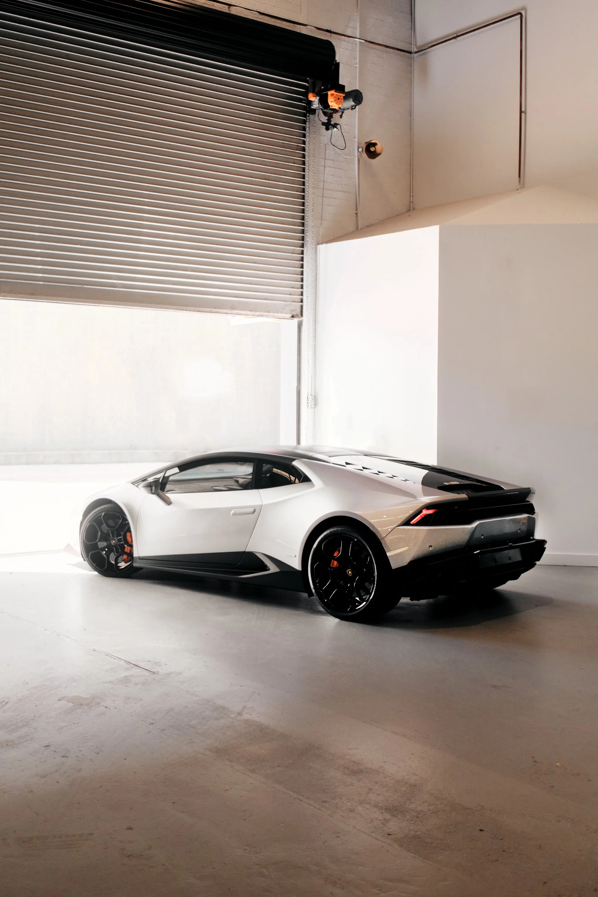 A silver Lamborghini sports car parked inside a minimalistic, industrial-style garage with white walls and a concrete floor. The car has black rims with orange brake calipers, and the garage features a large metal roll-up door and some visible electr