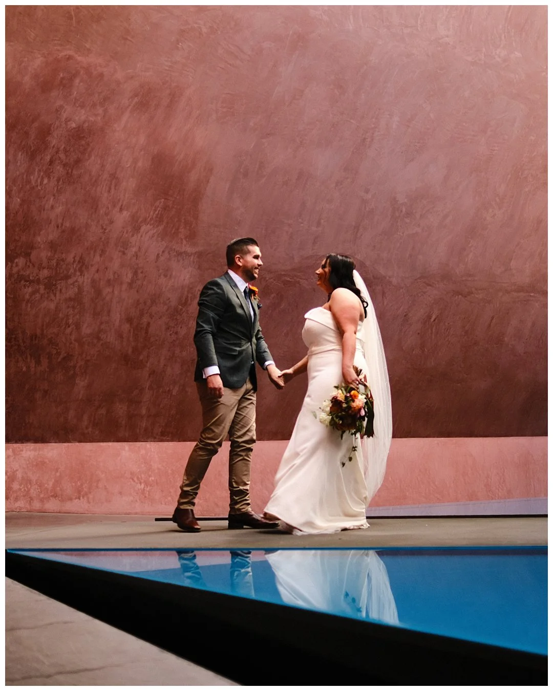 A couple gets married near a pool, holding hands, with a plain pink wall in the background.
