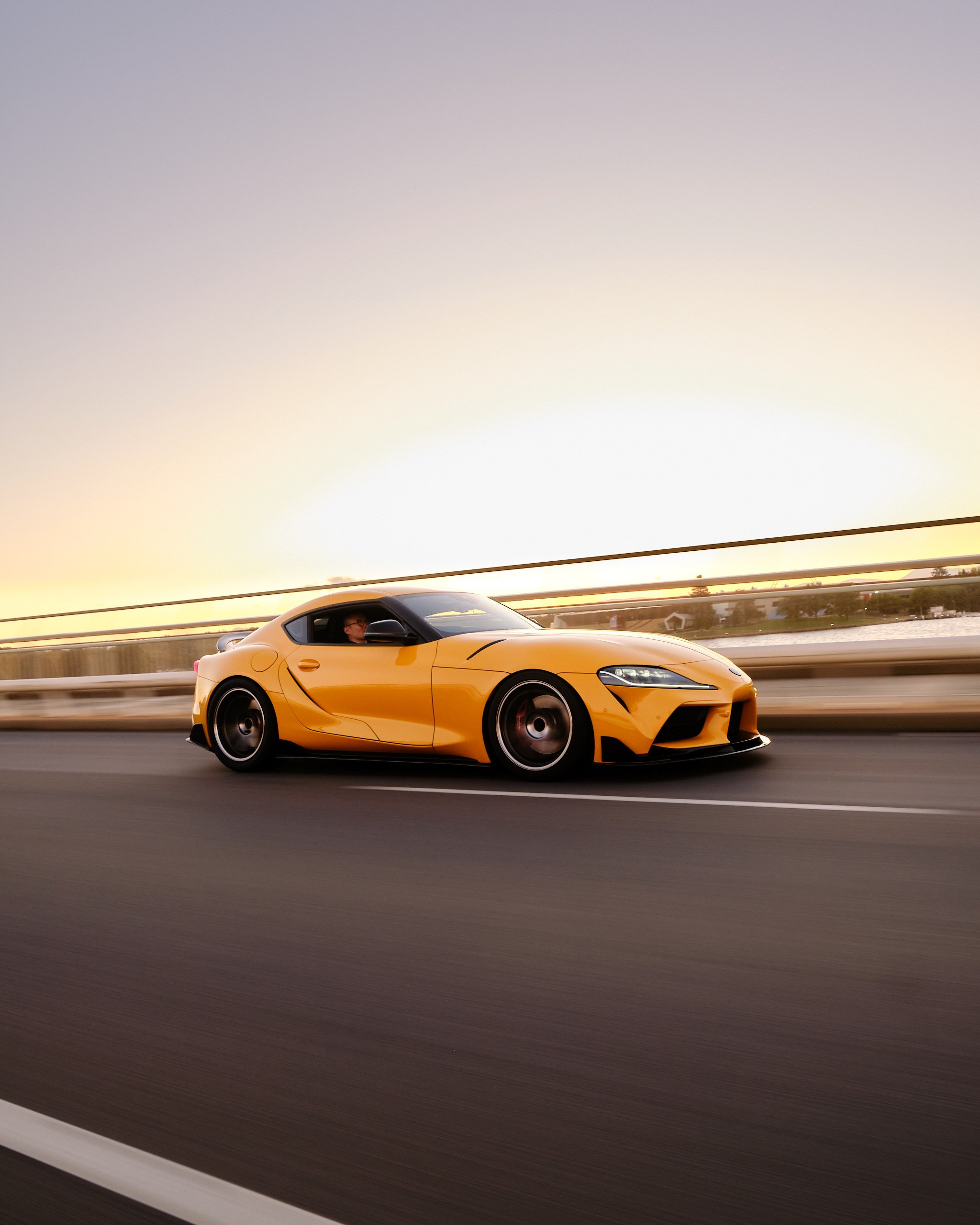 A yellow sports car driving on a highway at sunset, with a person visible in the driver’s seat and a guardrail along the side of the road.