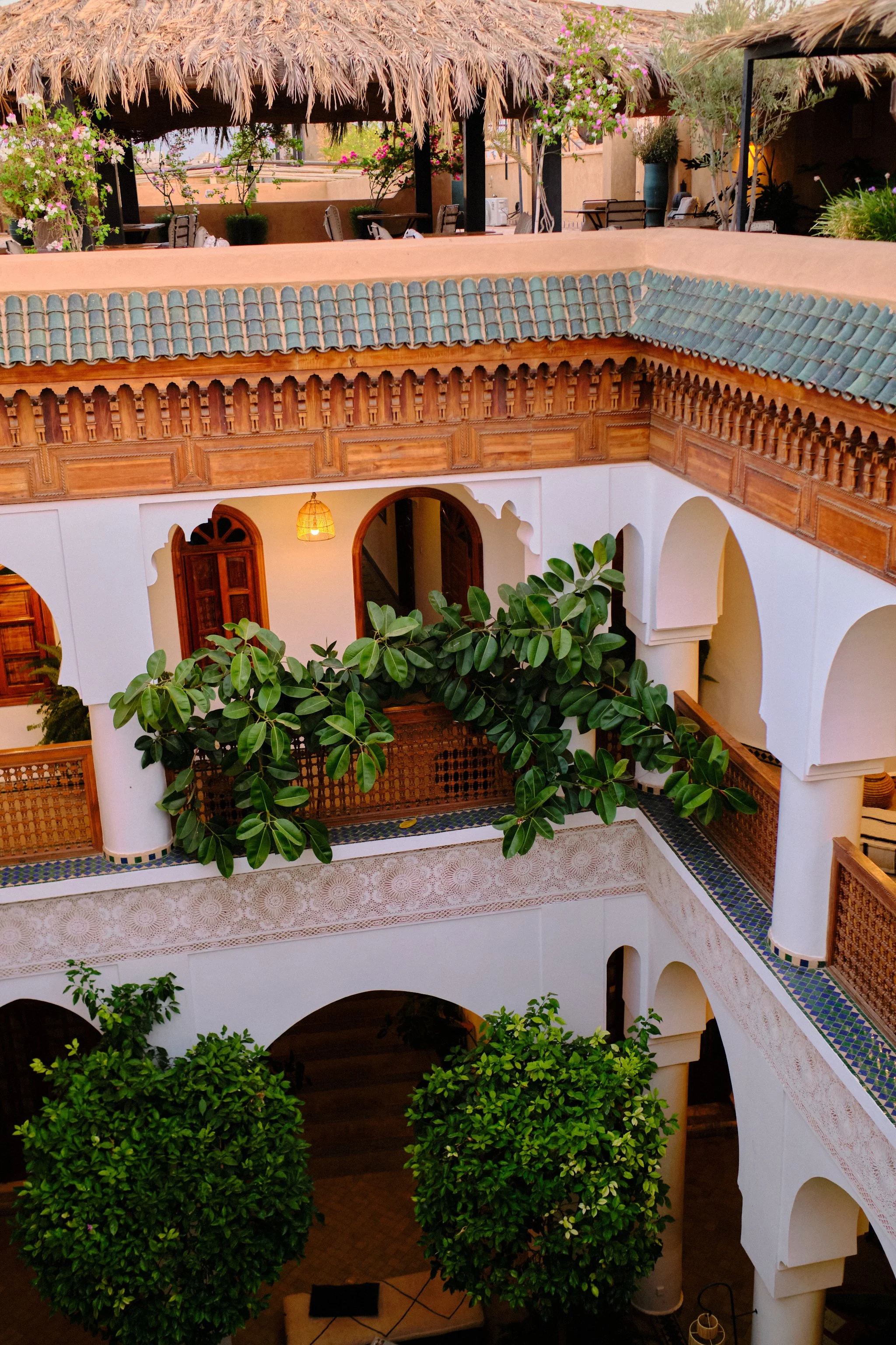 Interior view of a traditional courtyard with white walls, arched doorways, greenery, and a second-story balcony with decorative woodwork and tiles, glimpse of a hanging lantern, and plants with pink flowers.