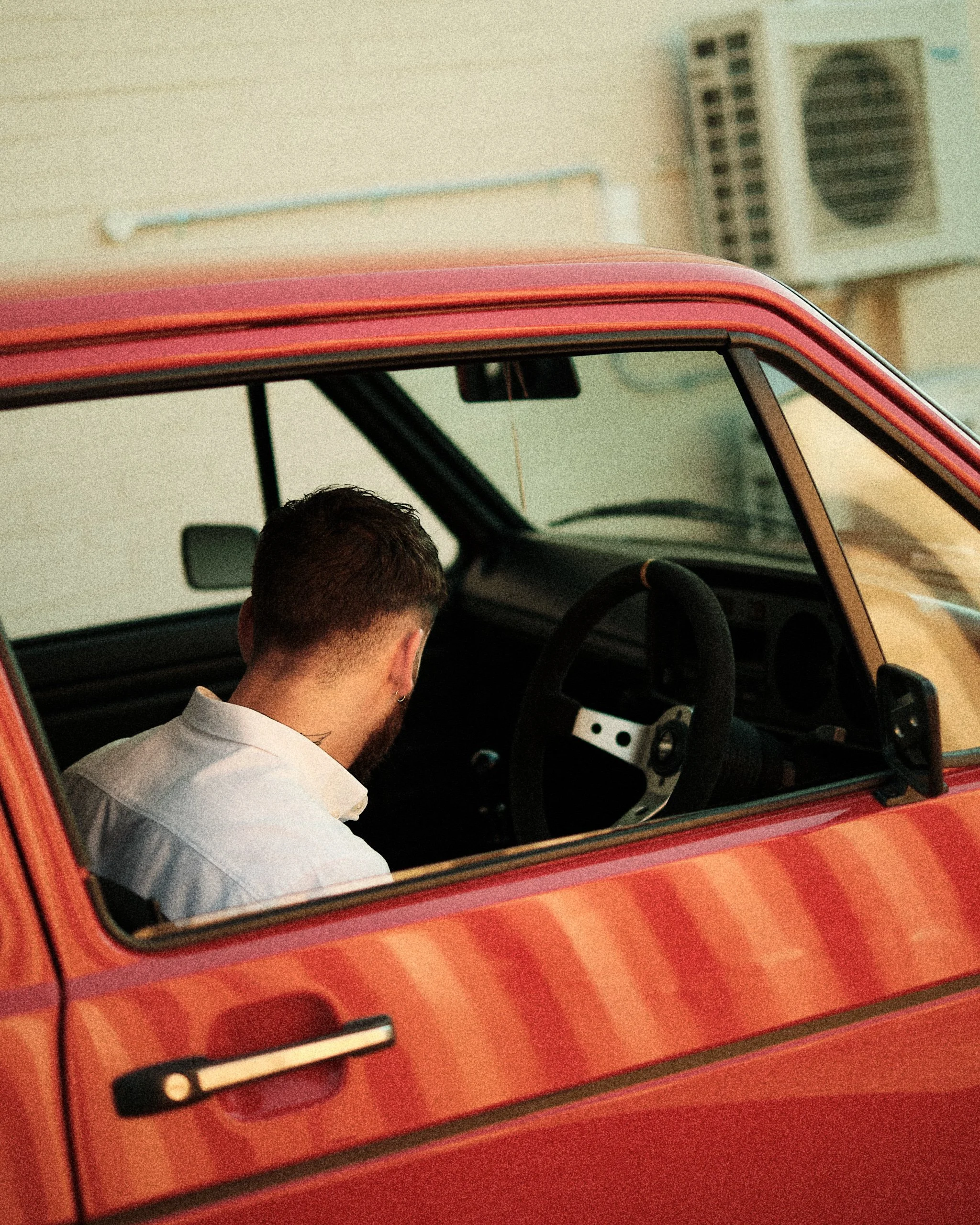 Rear view of a person sitting inside a red vintage car with a focus on the head and upper shoulders, visible through the car window, with the background showing a building and air conditioning units.