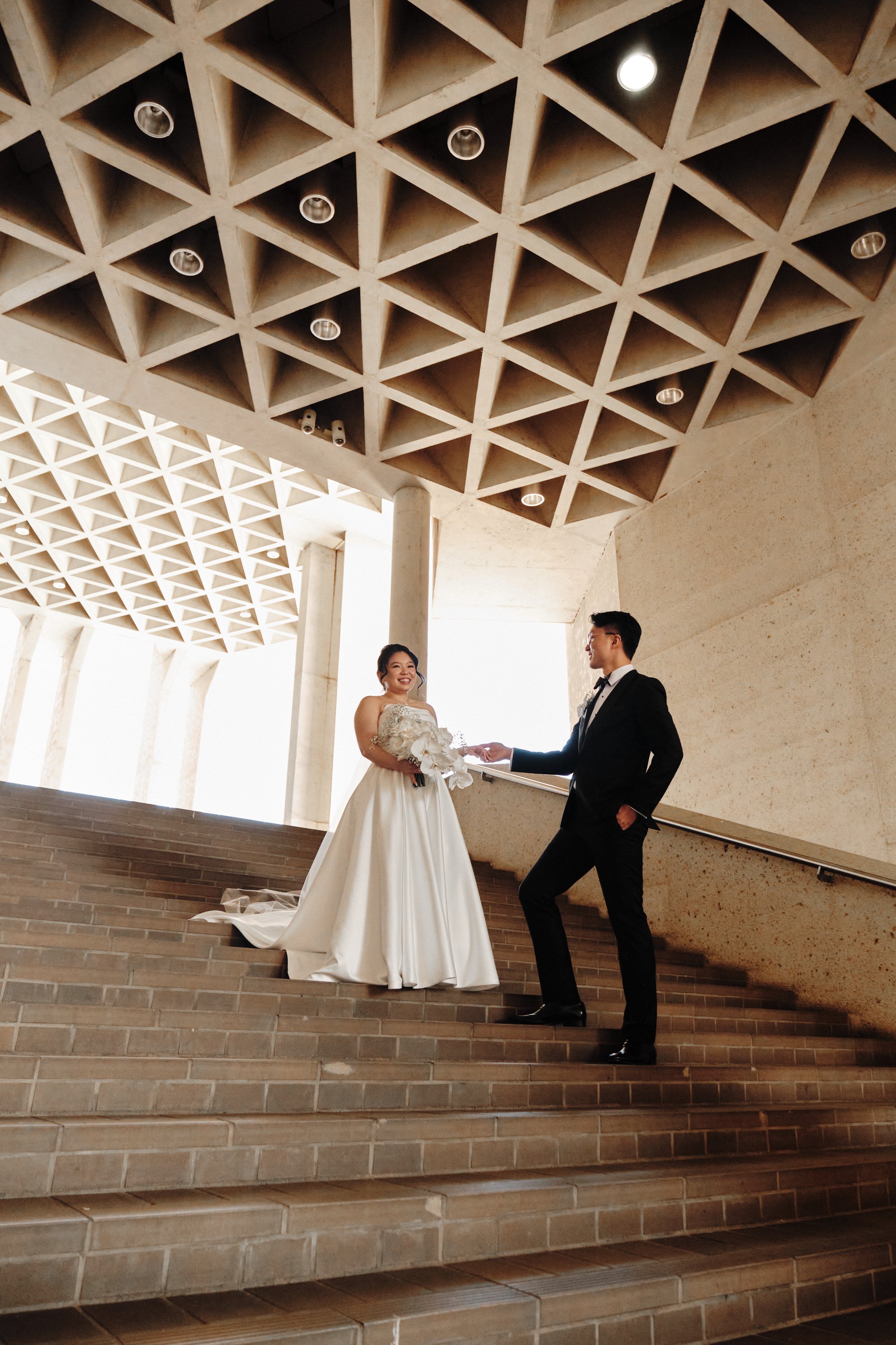 A bride and groom standing on steps inside a modern building with a grid-patterned ceiling, exchanging vows or rings, smiling at each other.